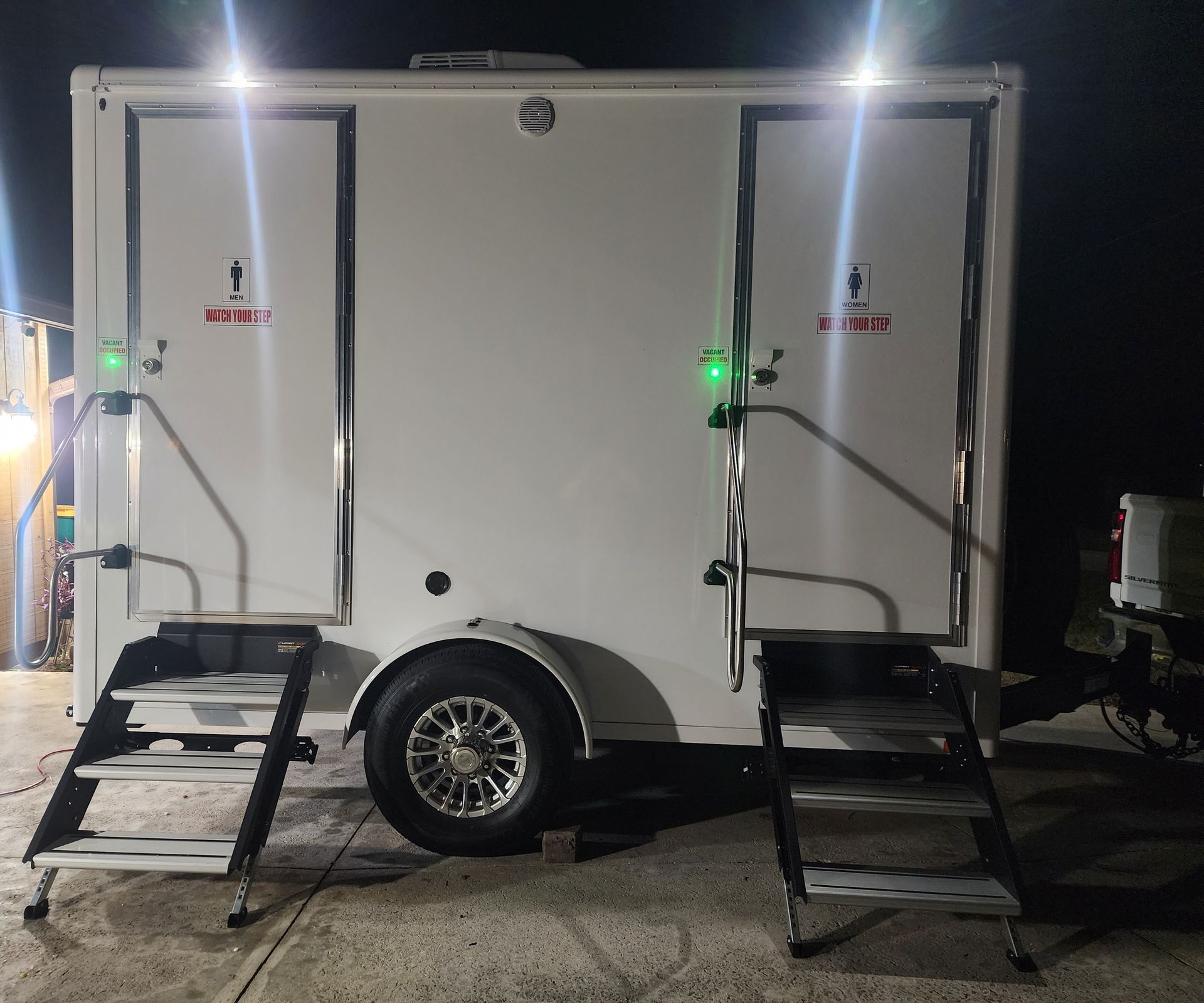 White enclosed trailer at night with two open side doors and stairs, lit by bright overhead lights.