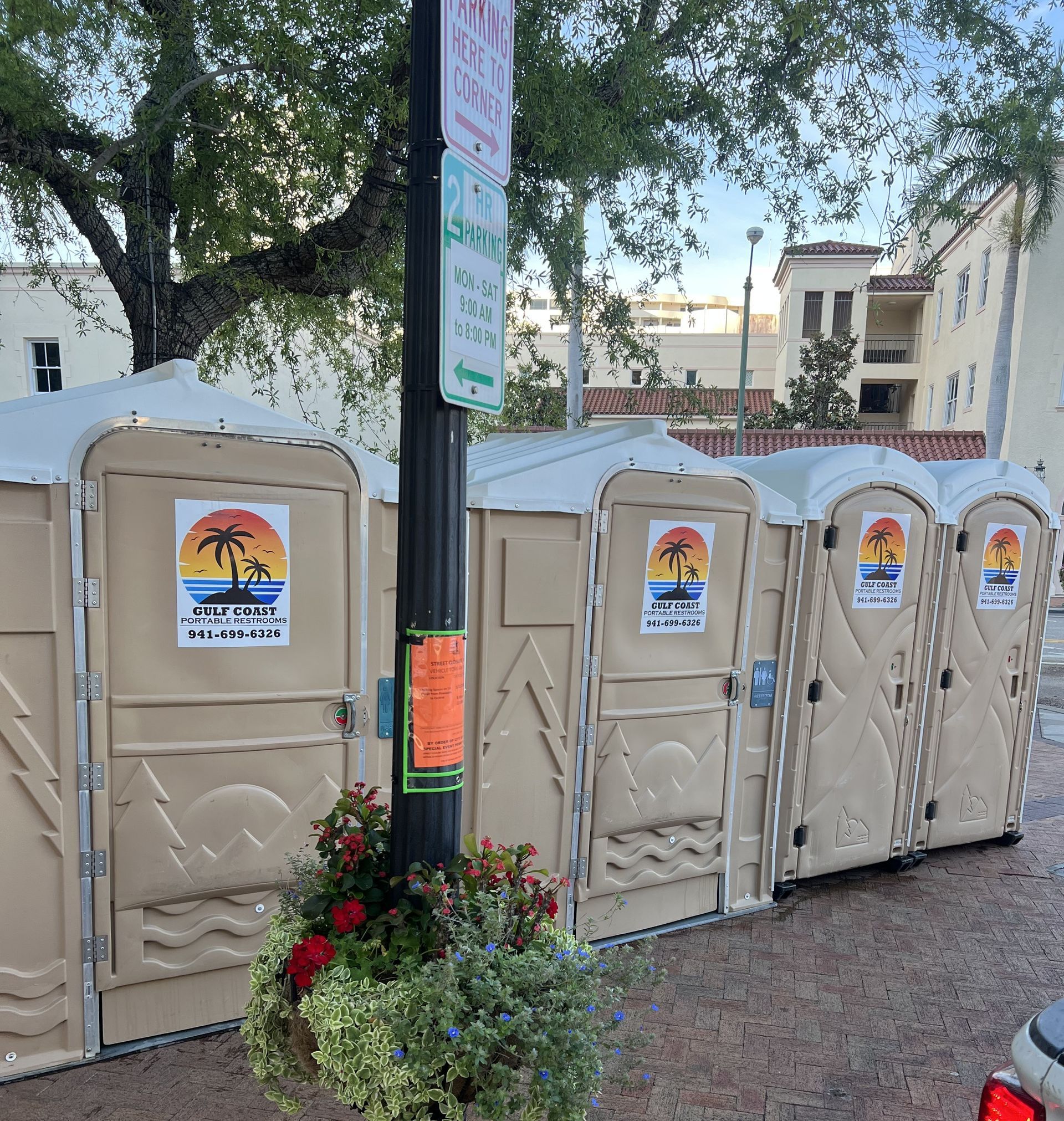 Row of beige portable toilets beside a pole and flower planter on a brick-paved street