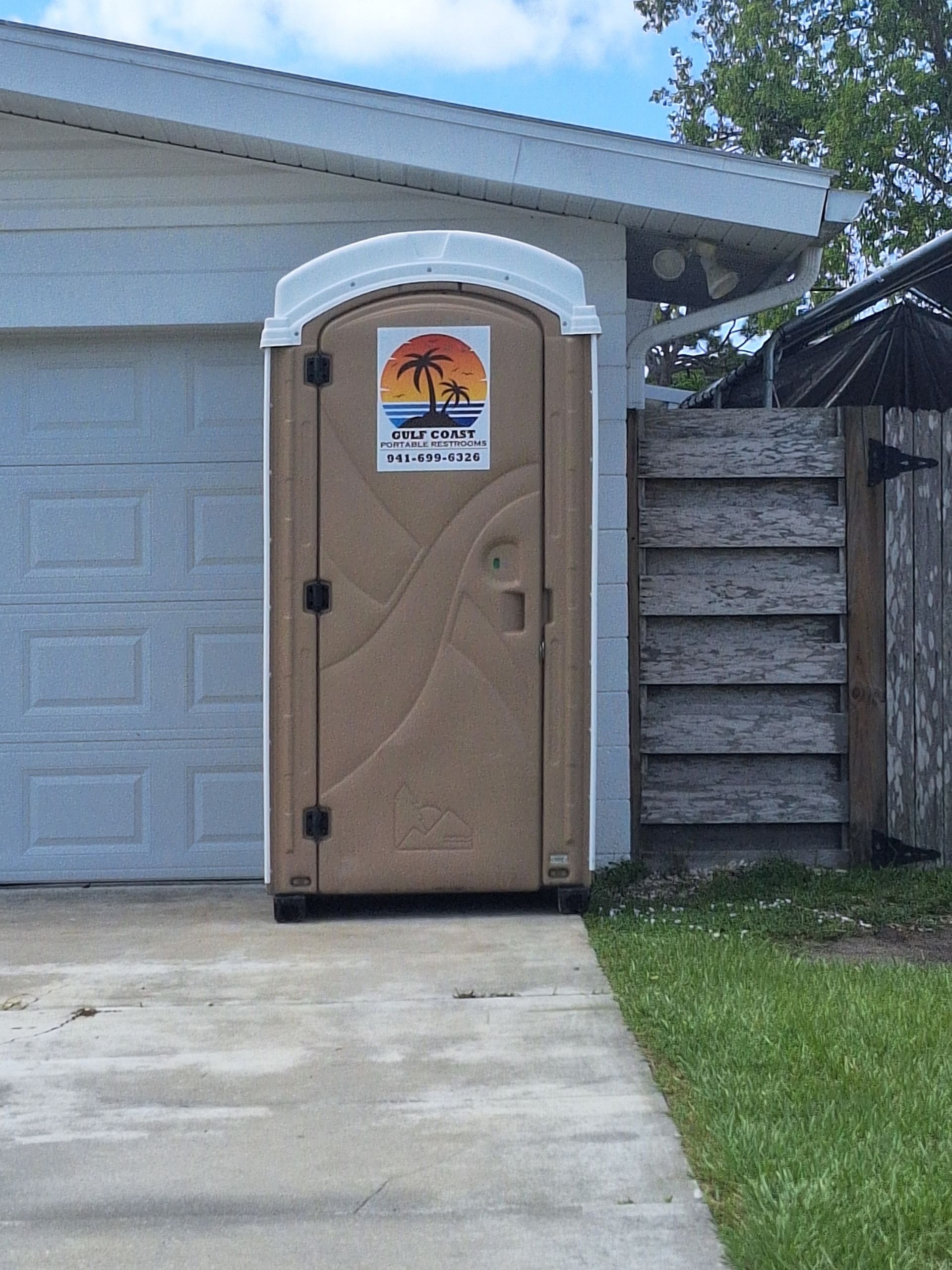 Tan portable toilet beside a garage, with a palm-tree sign on the door.