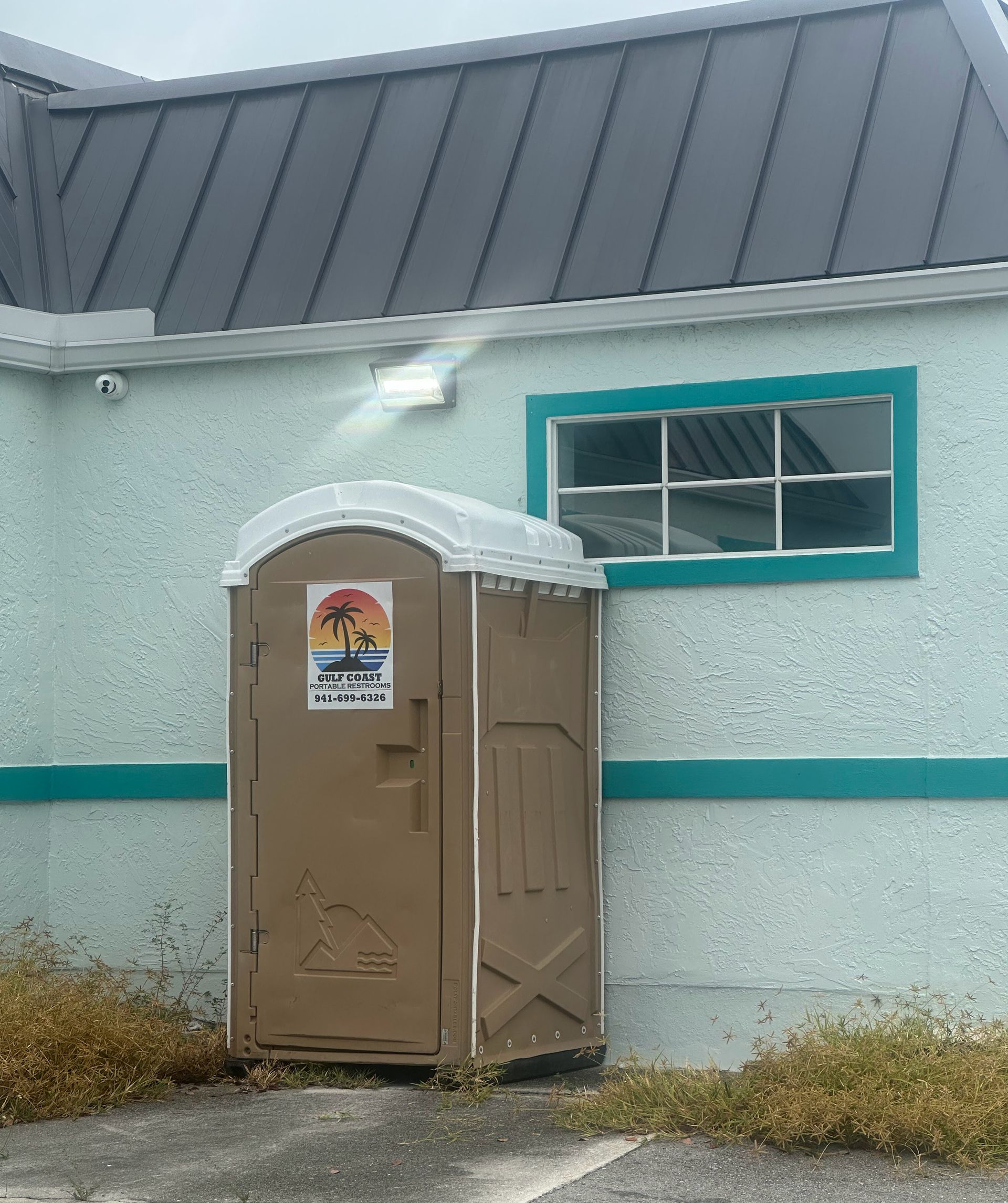 Brown portable toilet beside a pale green building with teal trim and a window