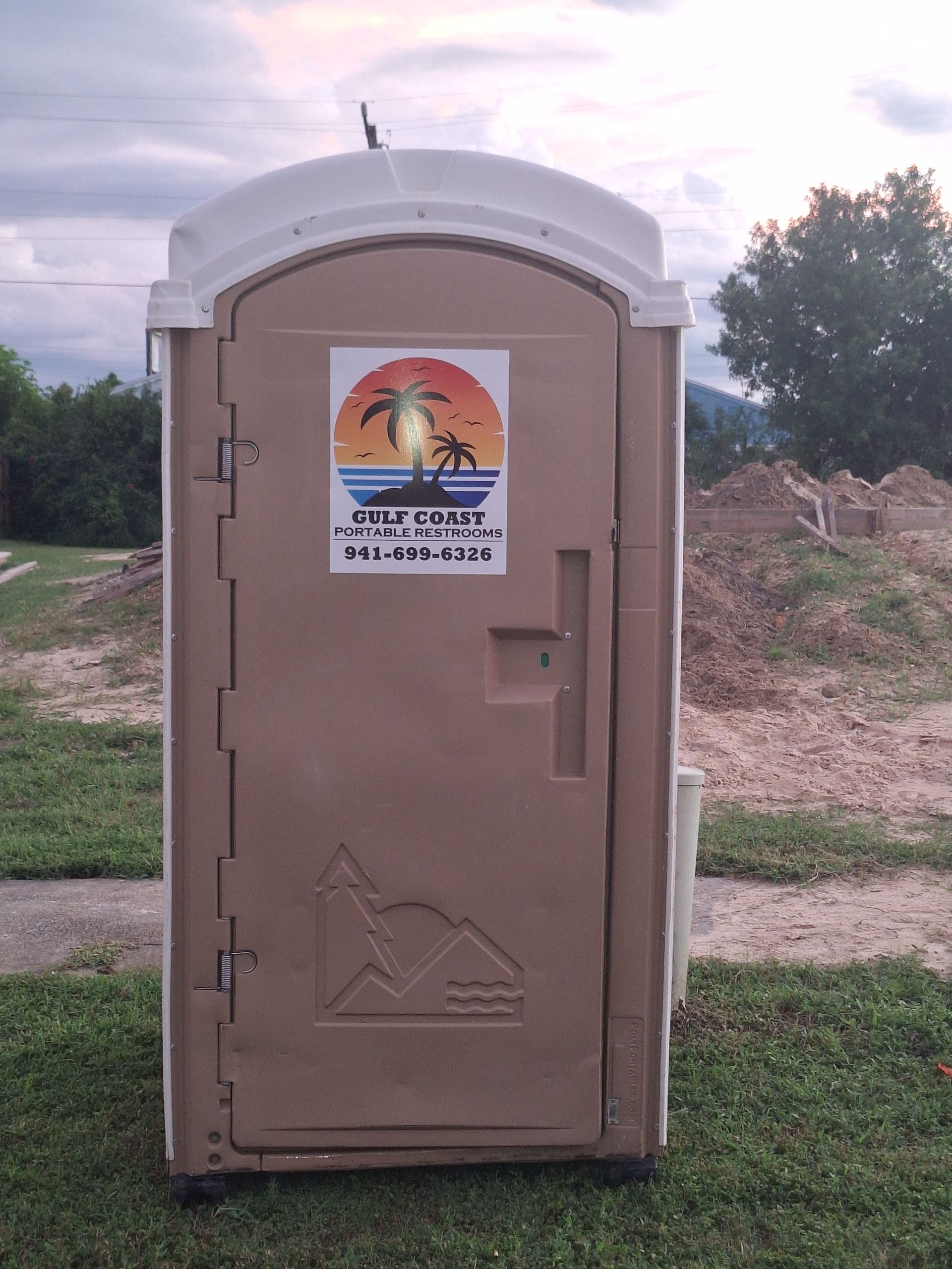 Brown portable toilet with a sunset sign, standing outdoors on grass near trees and dirt piles.
