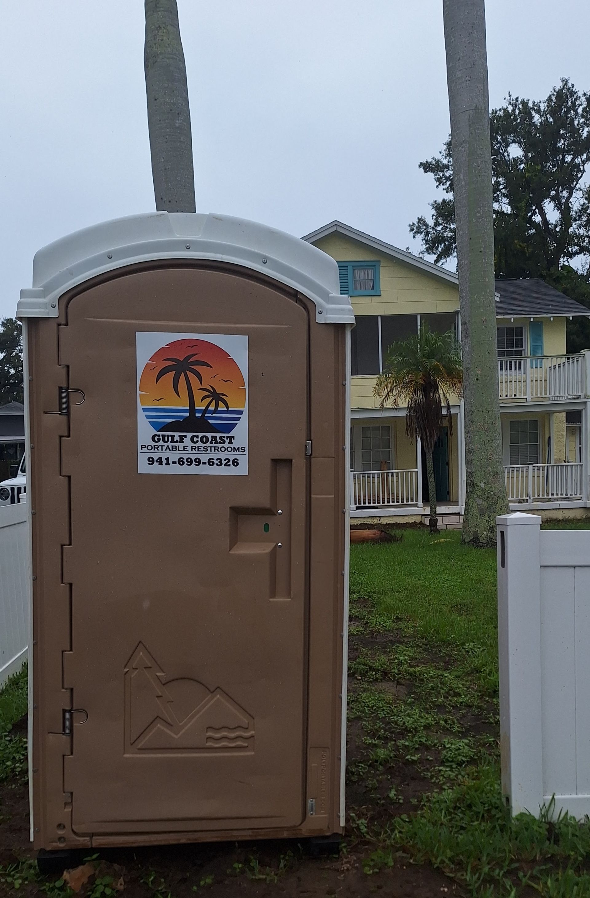 Tan portable toilet in front of a house, with a sunset beach sticker on the door.