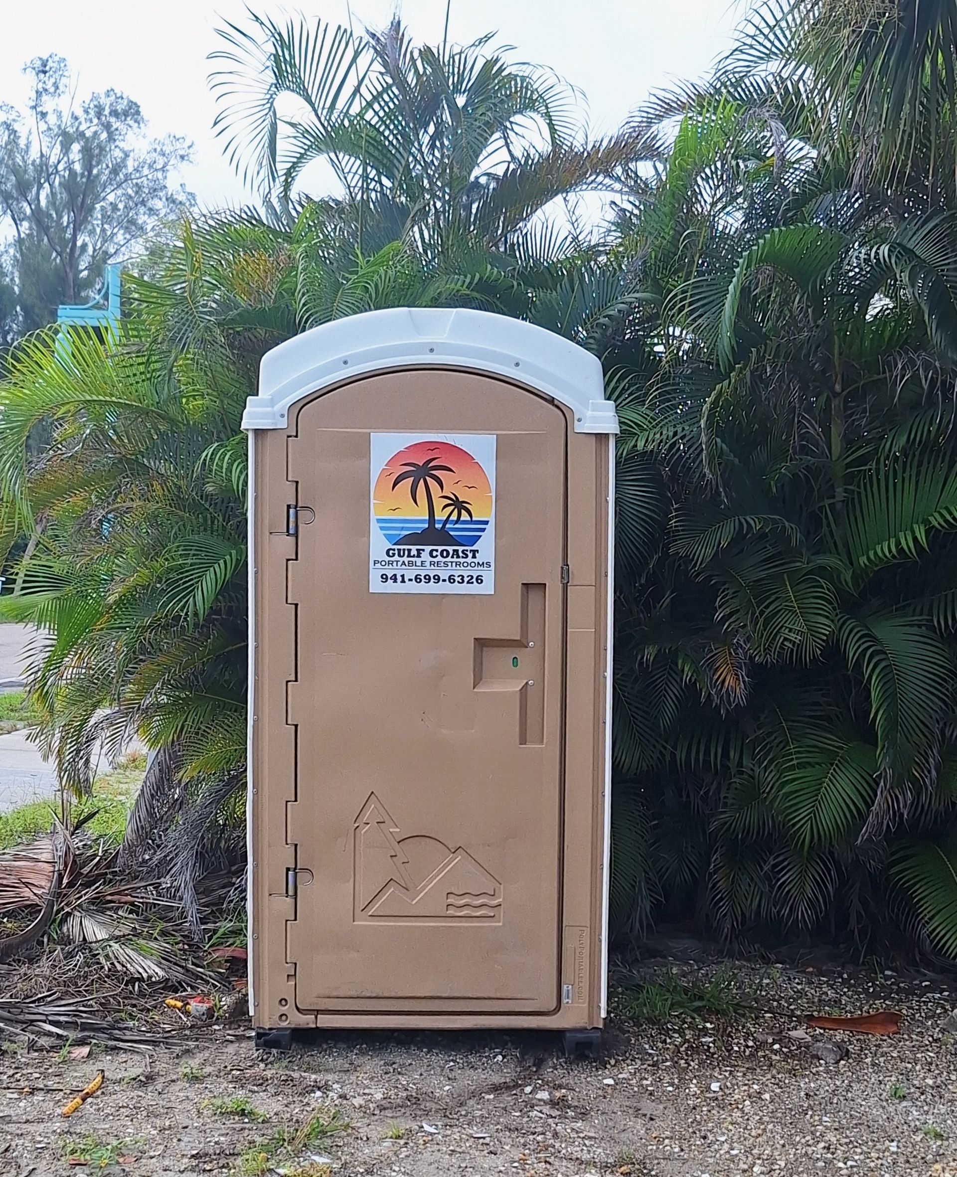Tan portable toilet with a sunset sign, set among trees and greenery outdoors.