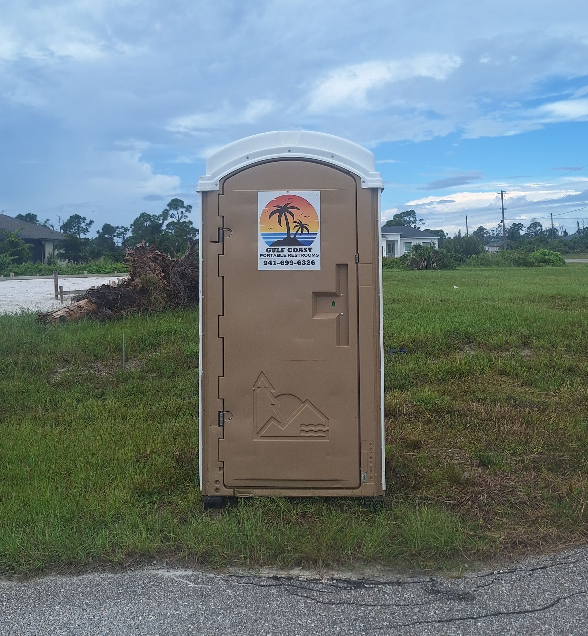 Tan portable toilet on a grassy roadside under a cloudy blue sky