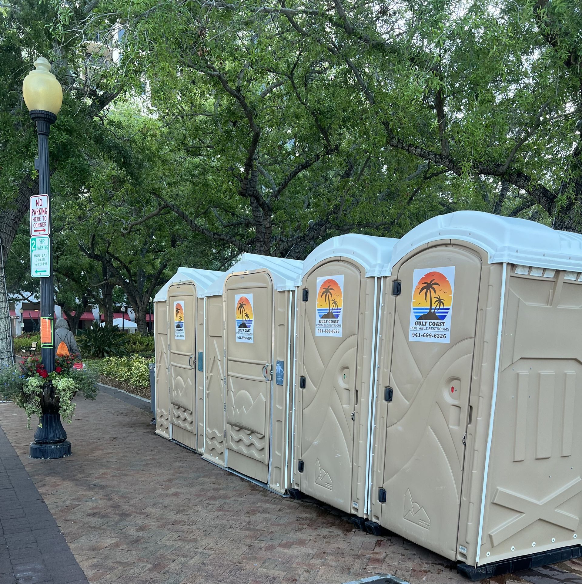 Row of beige portable toilets along a brick path beside trees and a lamppost in a park
