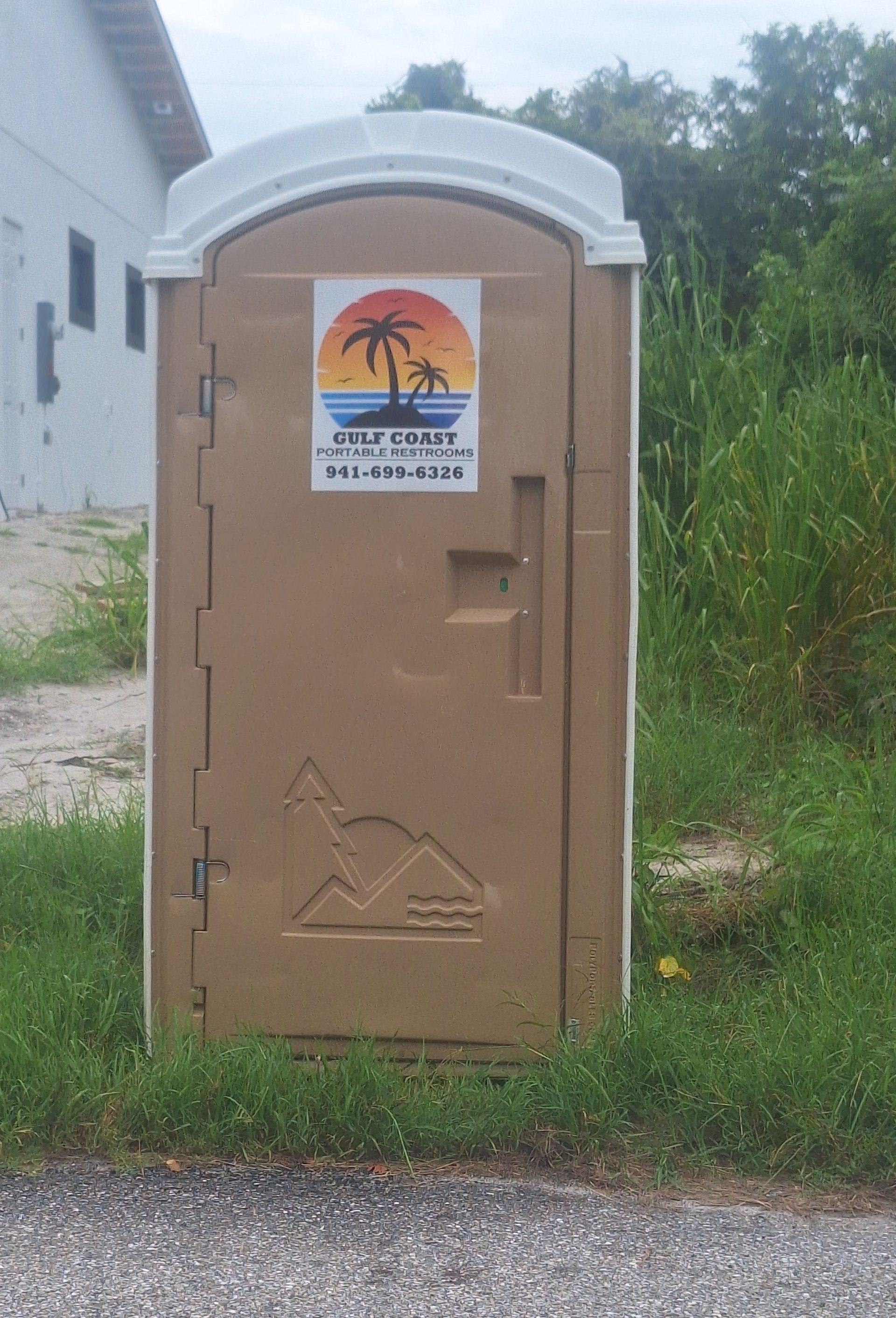 Tan portable restroom beside a white building, with a sunset sign on the door and grass around it
