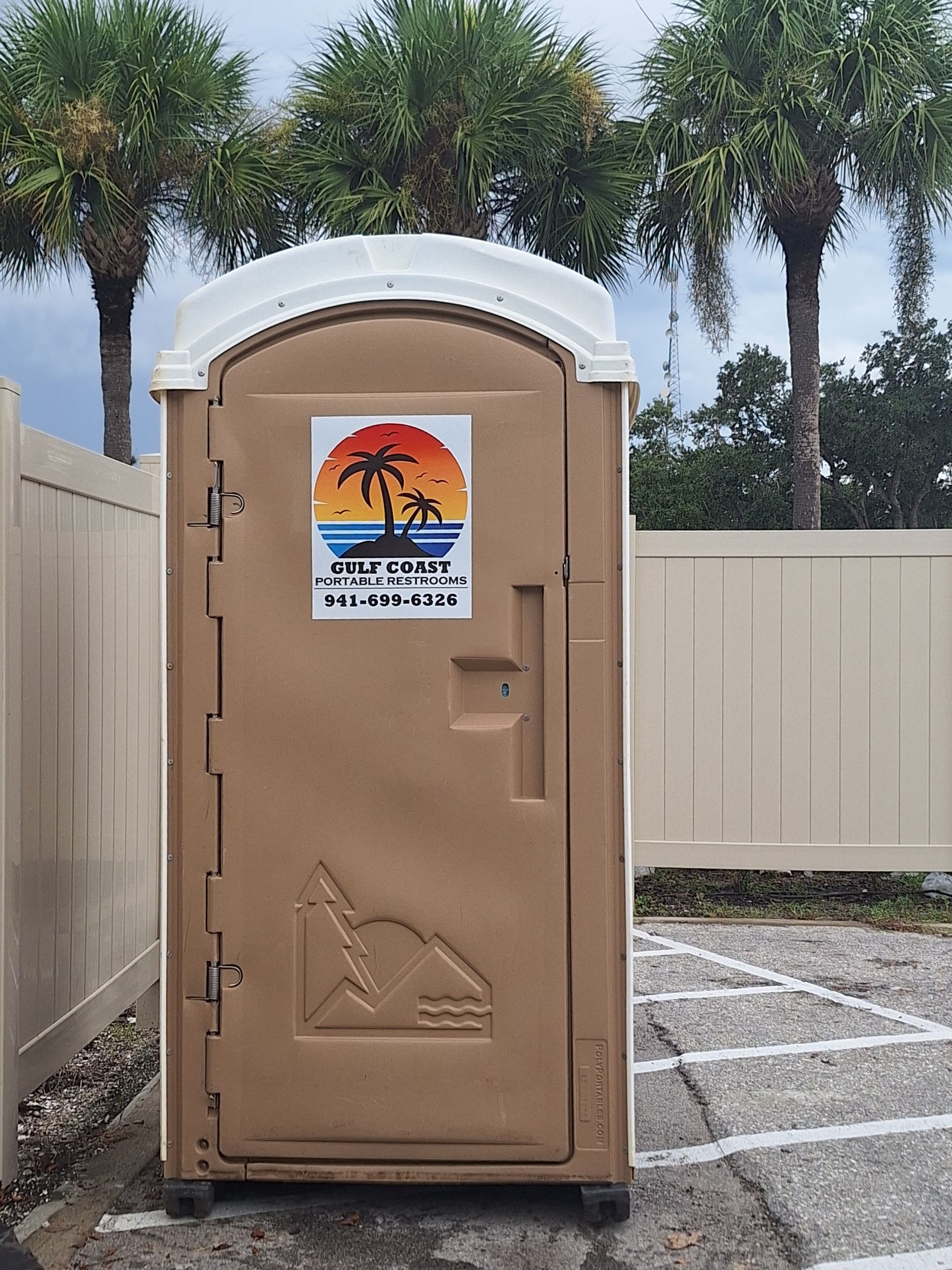 Tan portable restroom with beach sticker in a fenced outdoor lot, palm trees in background