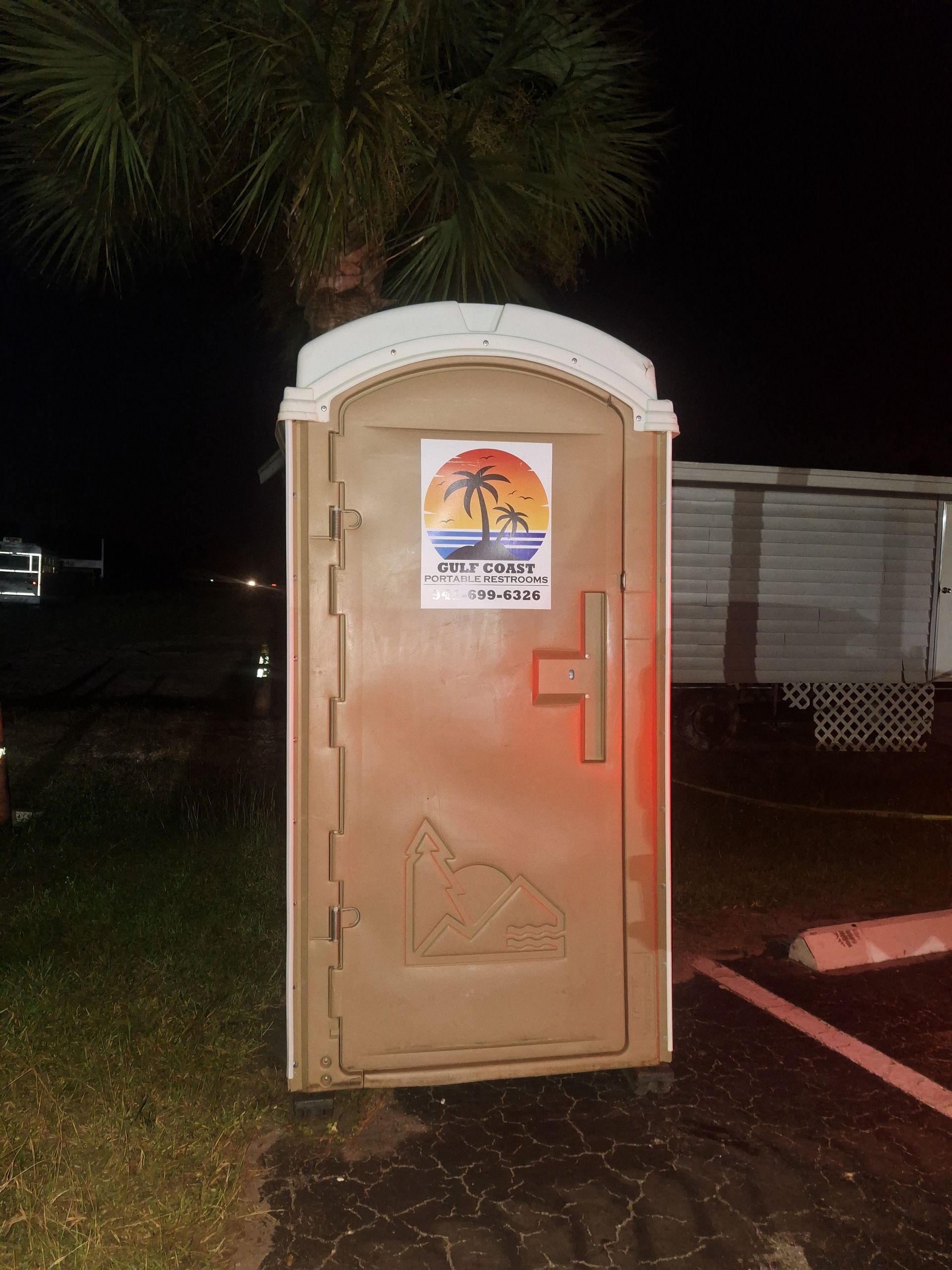 Portable toilet at night with a sign on the door, lit by warm light near a parking area.