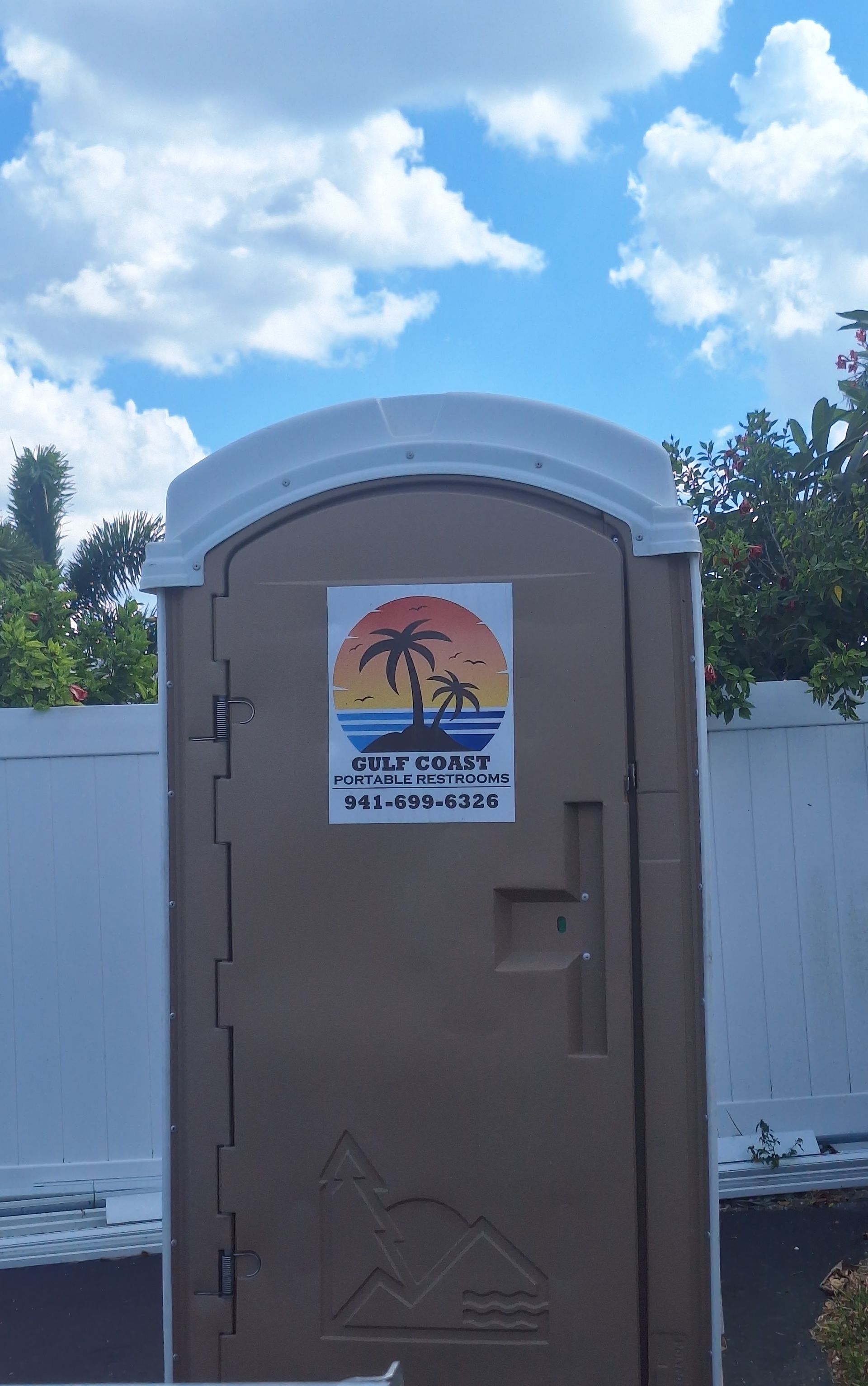 Portable restroom with a sunset beach sticker, standing outdoors against a white fence and blue sky.