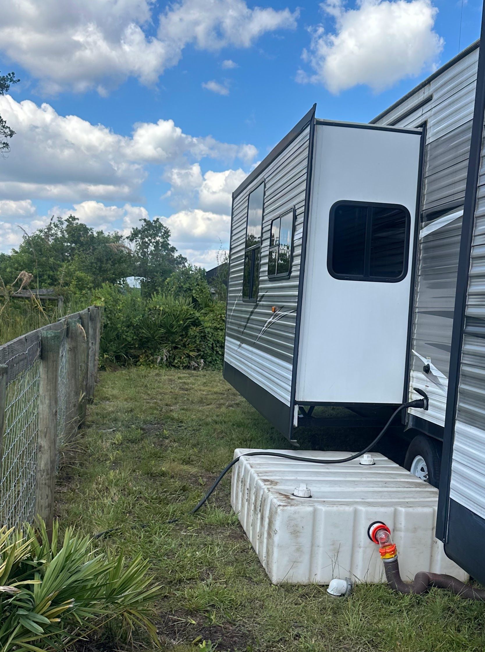 White trailer beside a fence with a white cooler and hose on grass under a cloudy sky