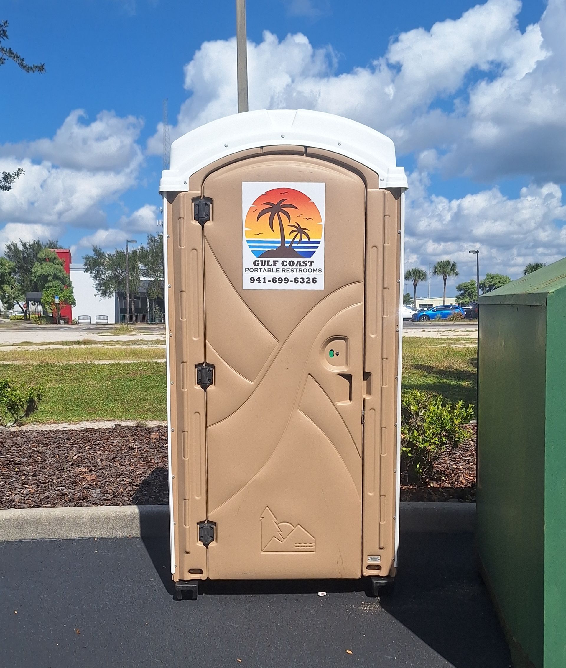 Beige portable toilet with a sunset poster, standing on pavement outdoors under a blue sky.