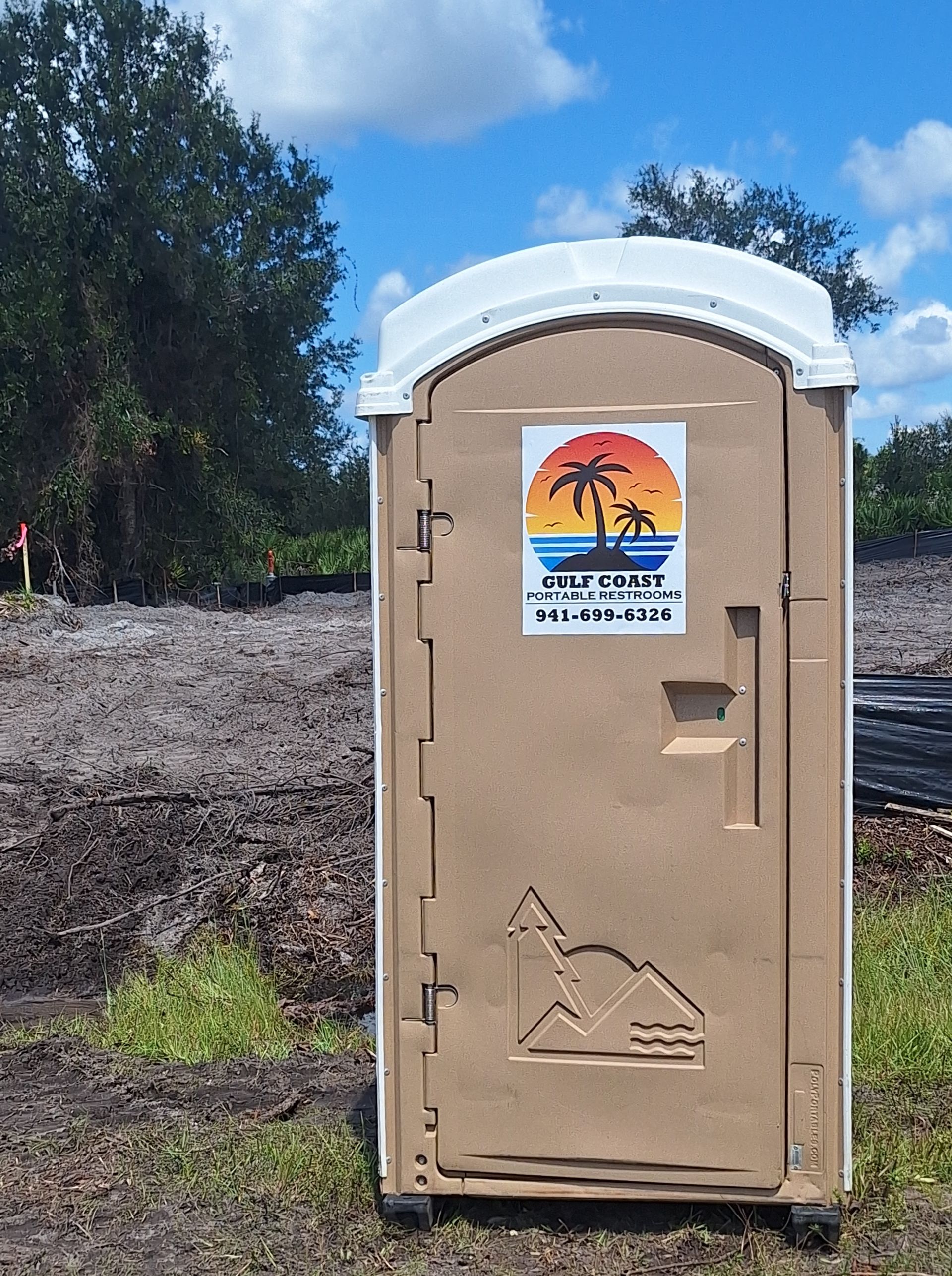 Tan portable toilet with a white roof on muddy ground near trees