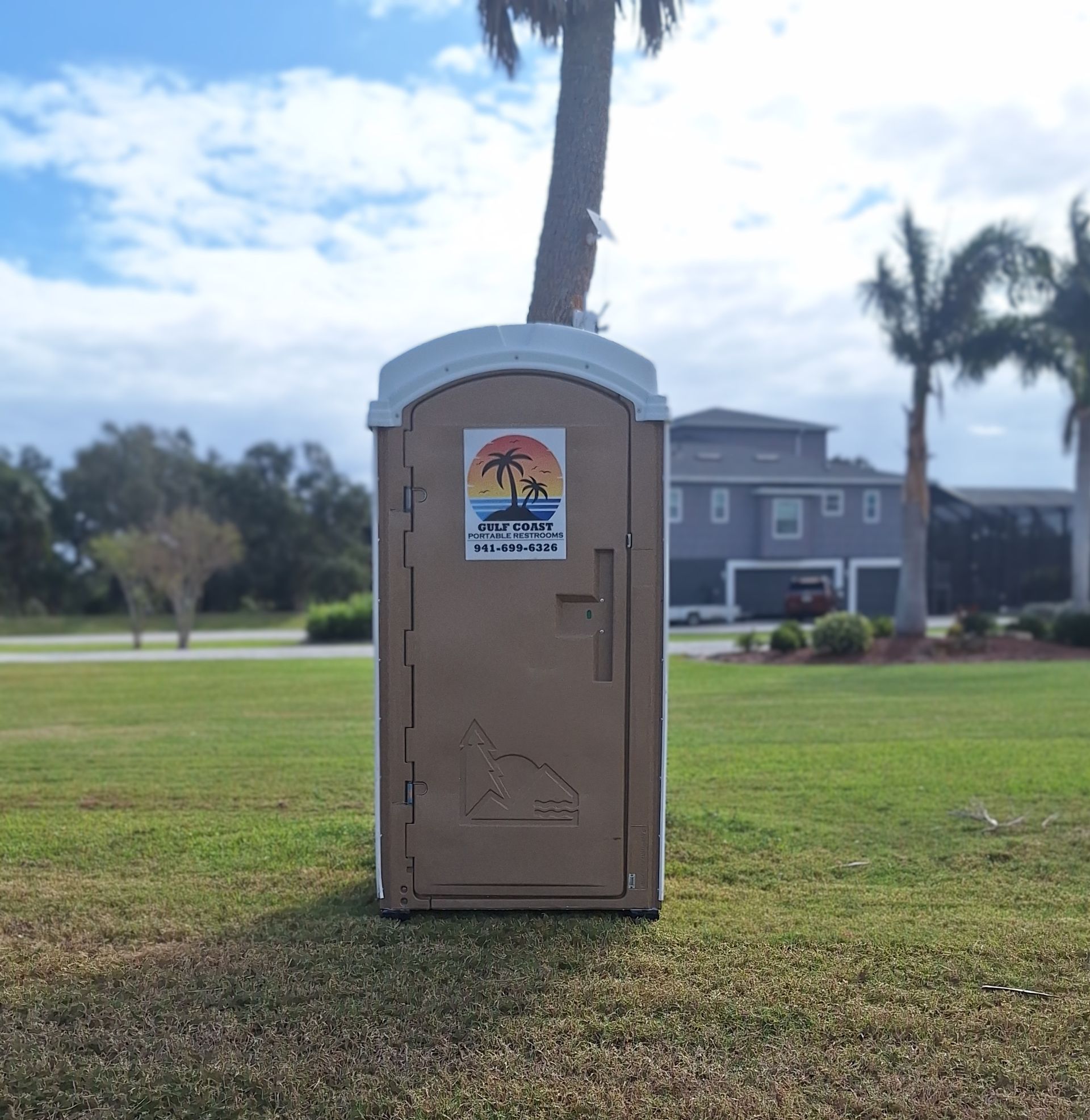 Portable toilet on a grassy field near a palm tree, with buildings and a cloudy sky in the background