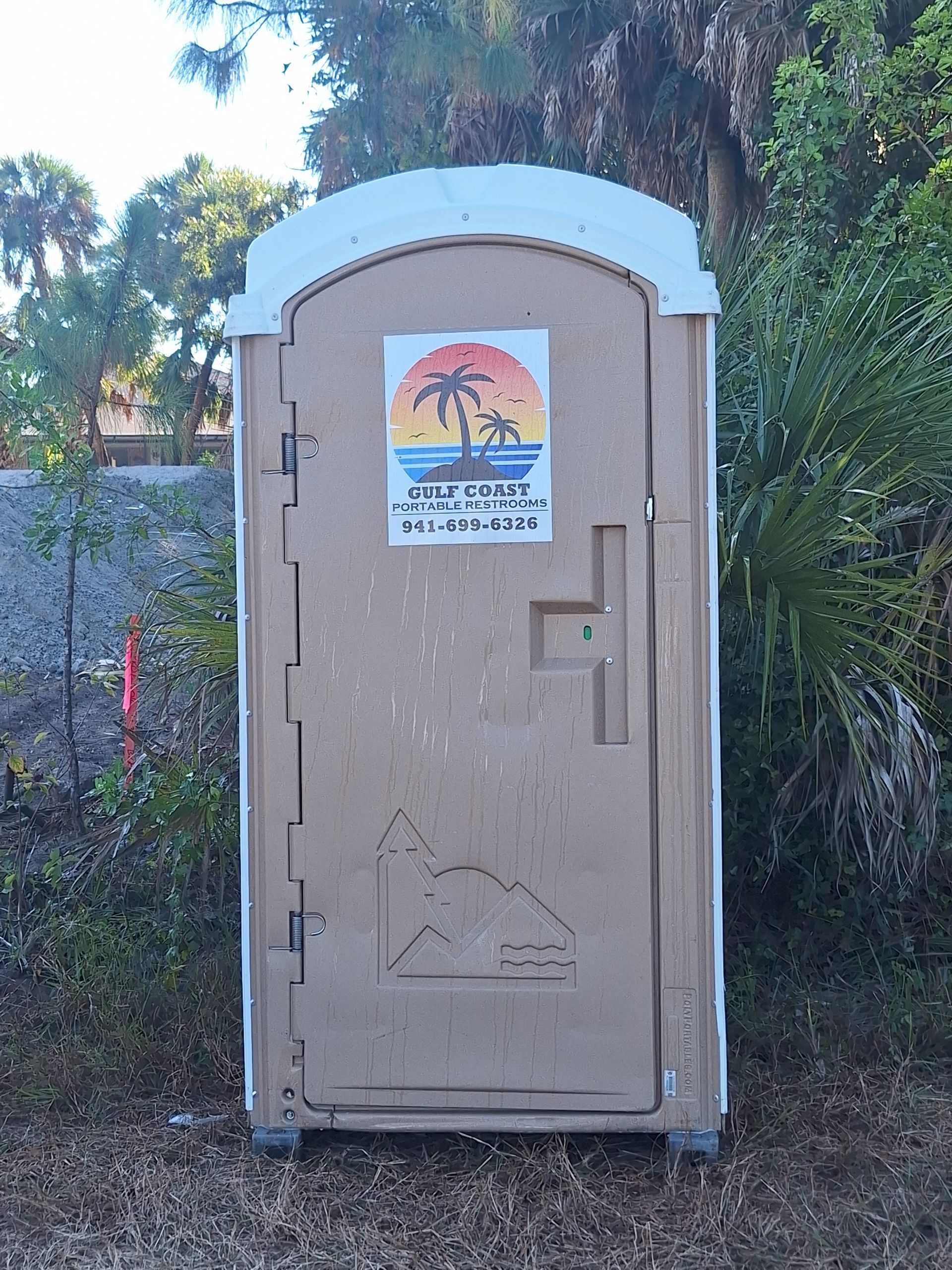 Tan portable toilet with a “Thankful” sign on the front, outdoors beside a fence and trees.