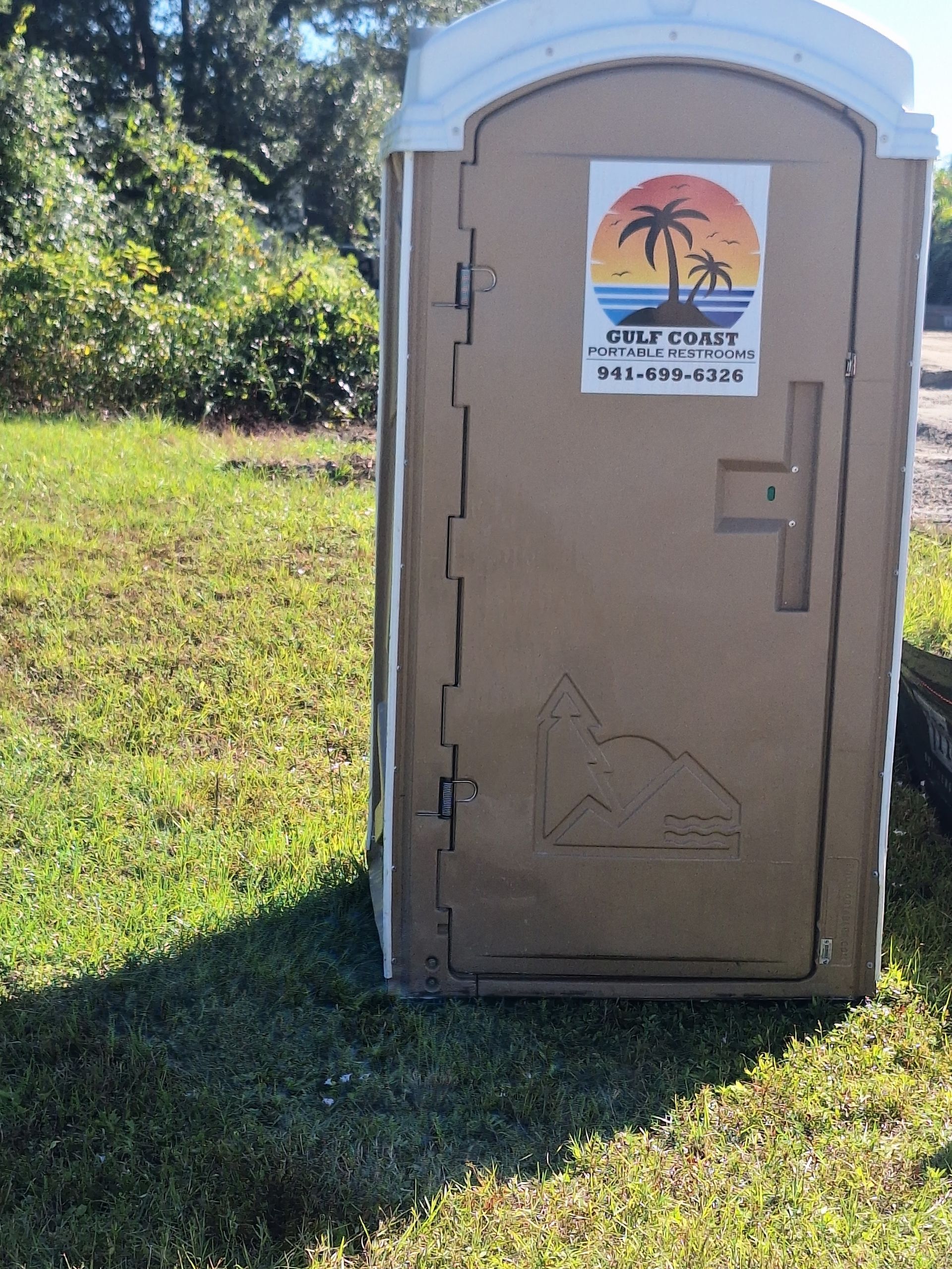 Portable toilet on grass with a sunset-themed sign on the door, outdoors near trees.