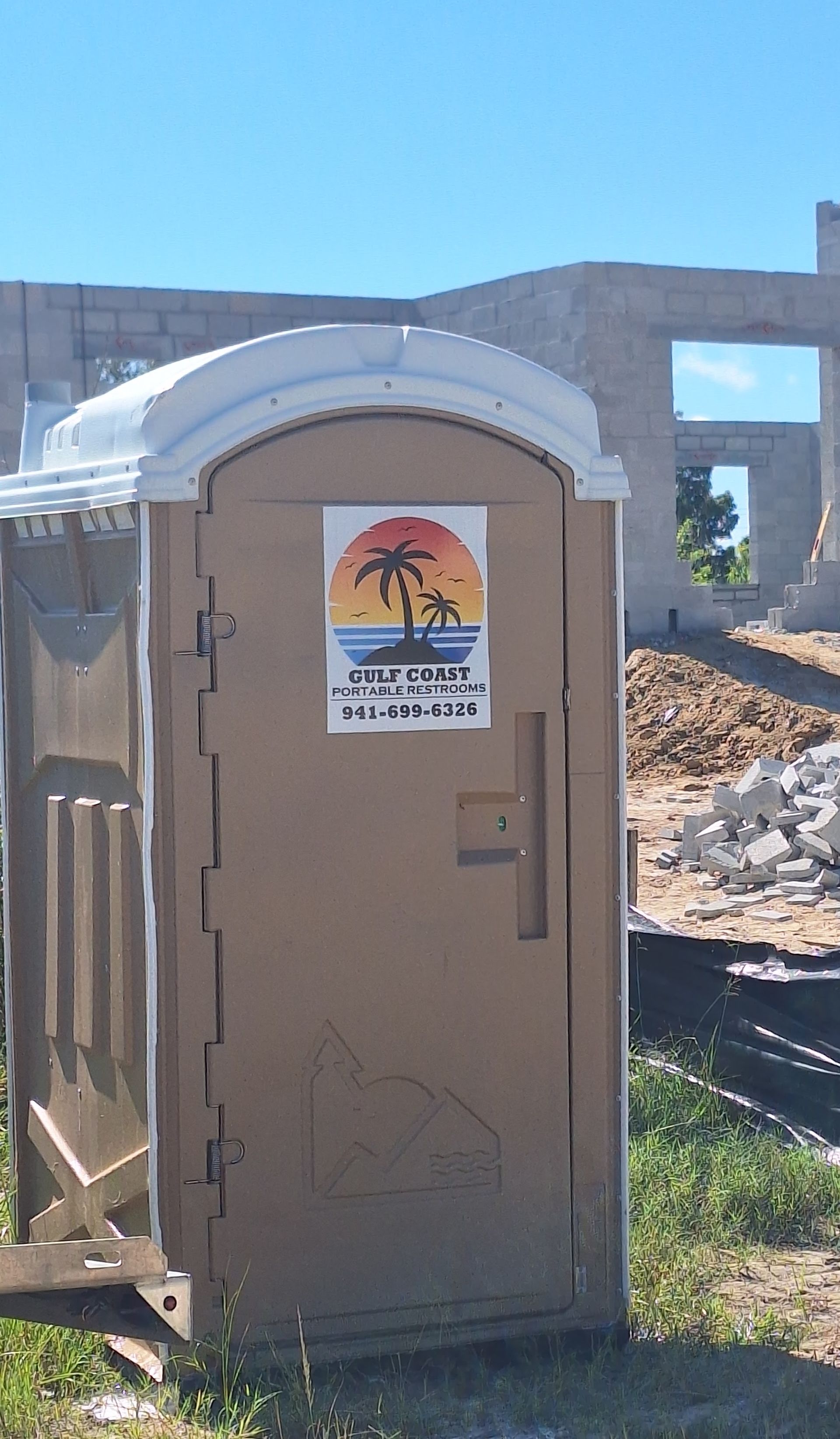 Portable toilet beside a cinderblock wall and rubble under a blue sky.