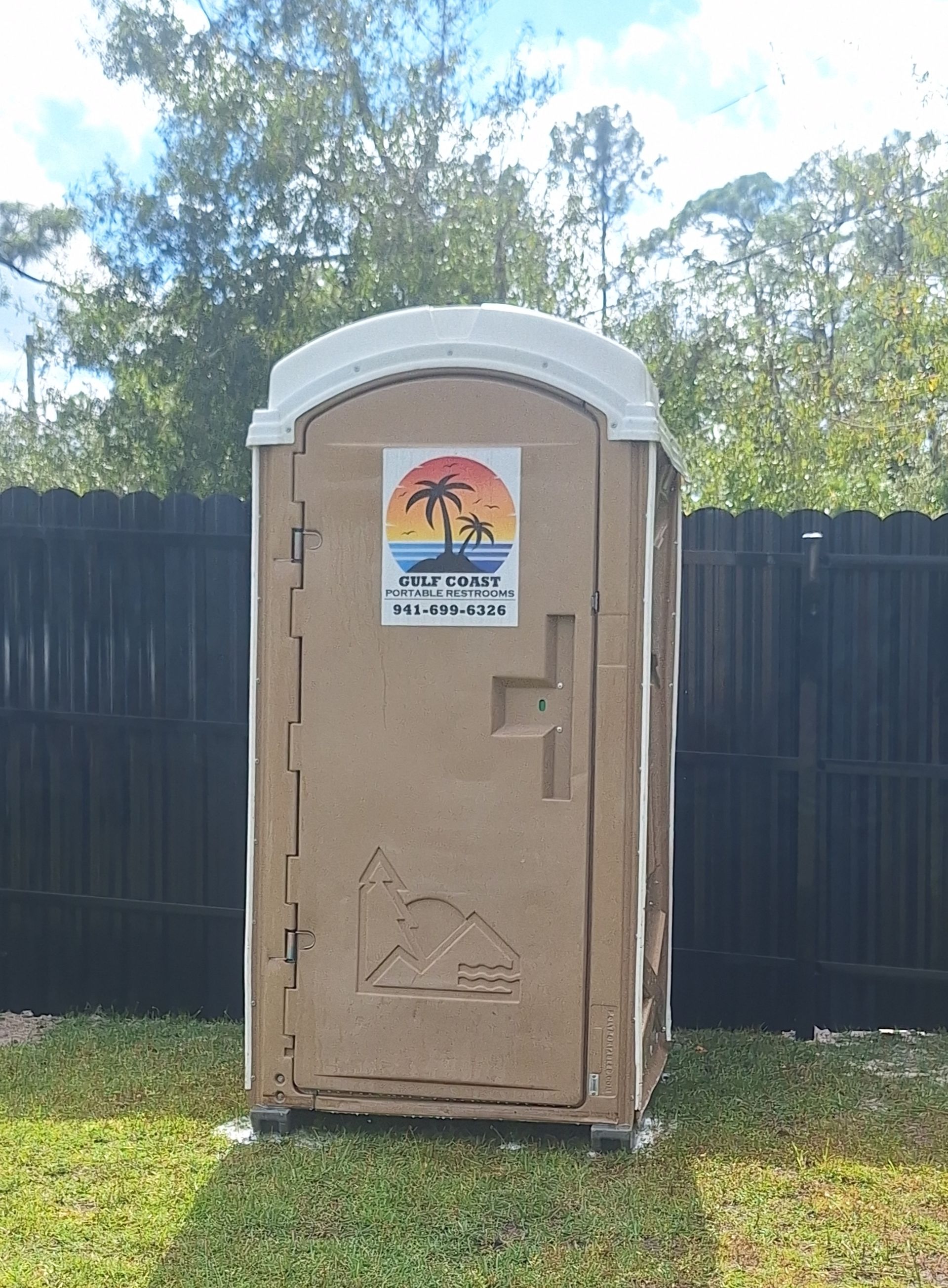 Tan portable toilet in a grassy yard with a black fence and trees behind it.
