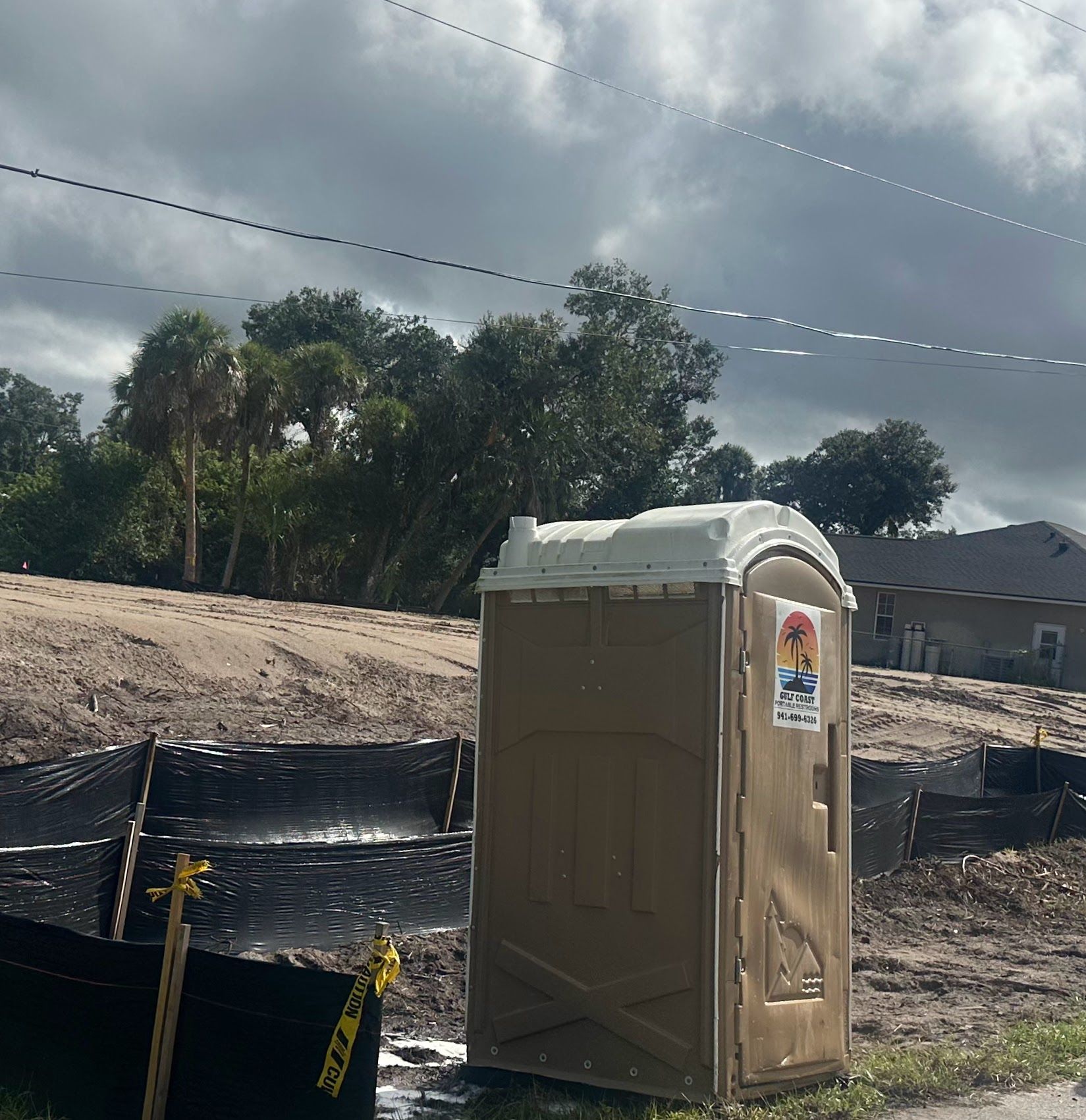 Tan portable toilet on a construction site with dirt, fencing, and cloudy sky.