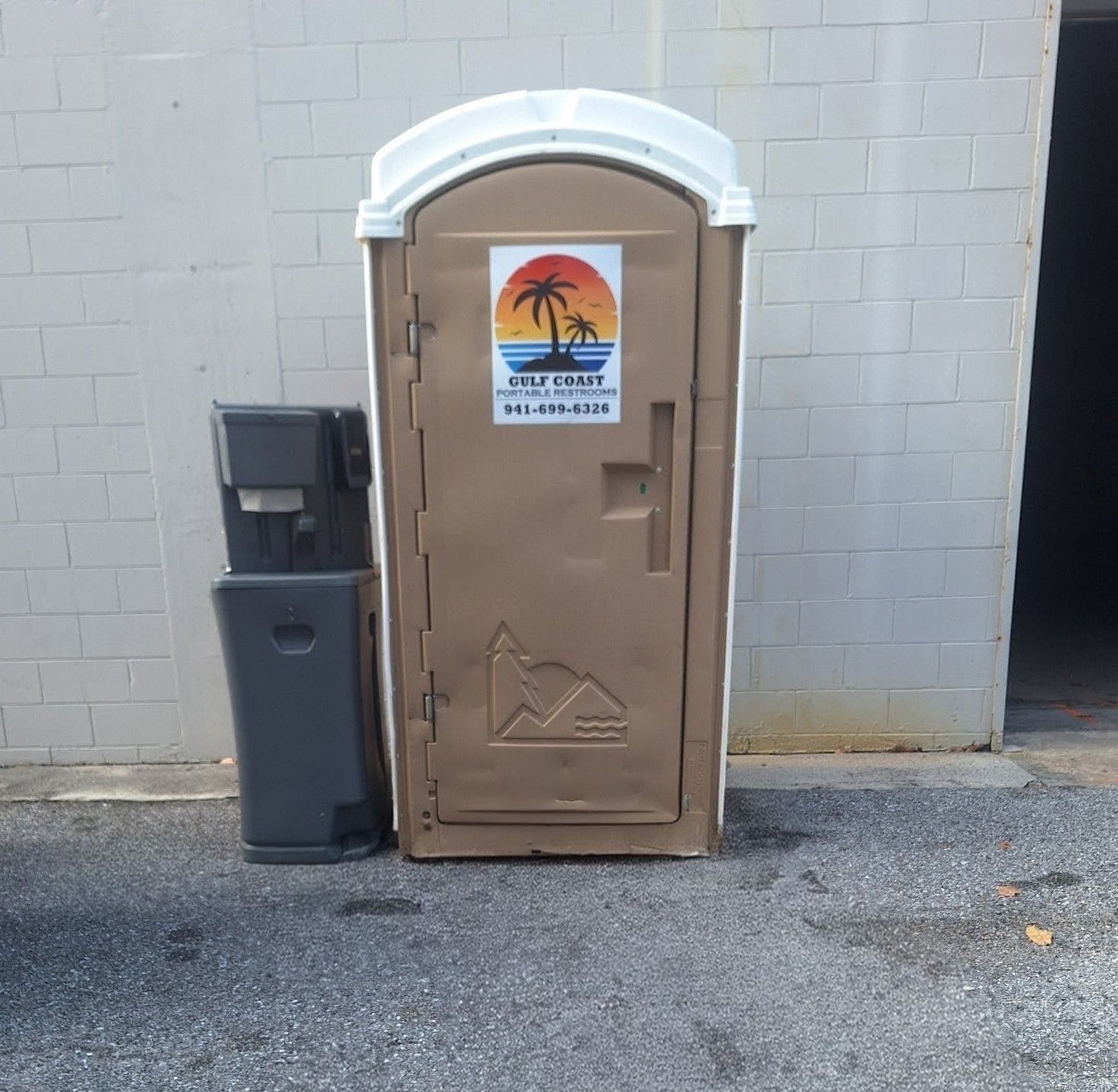 Portable tan restroom trailer beside a gray trash bin against a cinderblock wall.