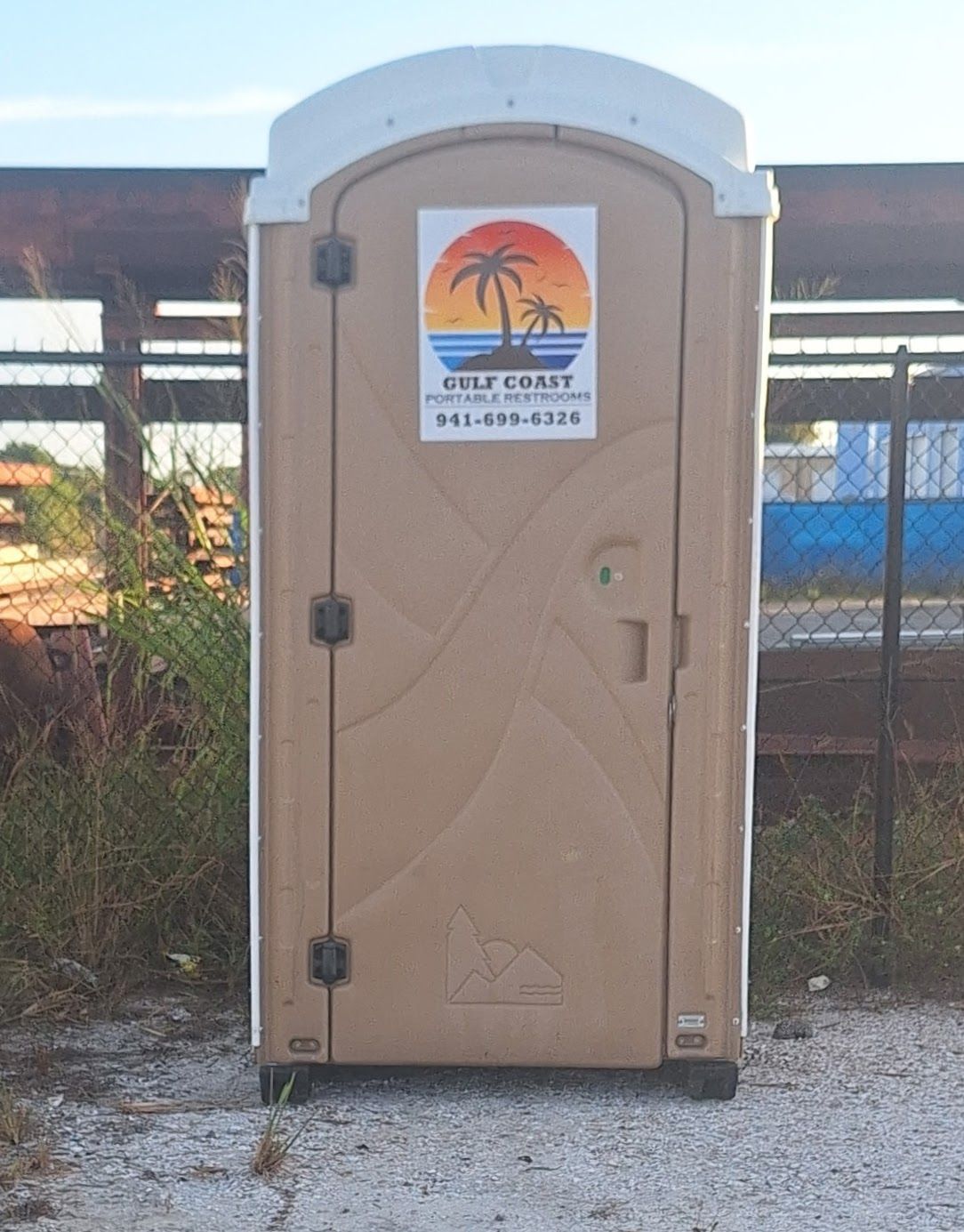 Tan portable toilet with a palm tree sign, standing on gravel outdoors near a fence.