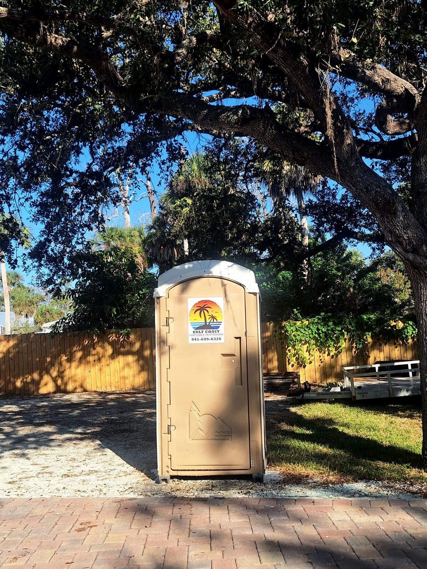 Tan portable restroom on a brick path beneath large trees in a sunny park area