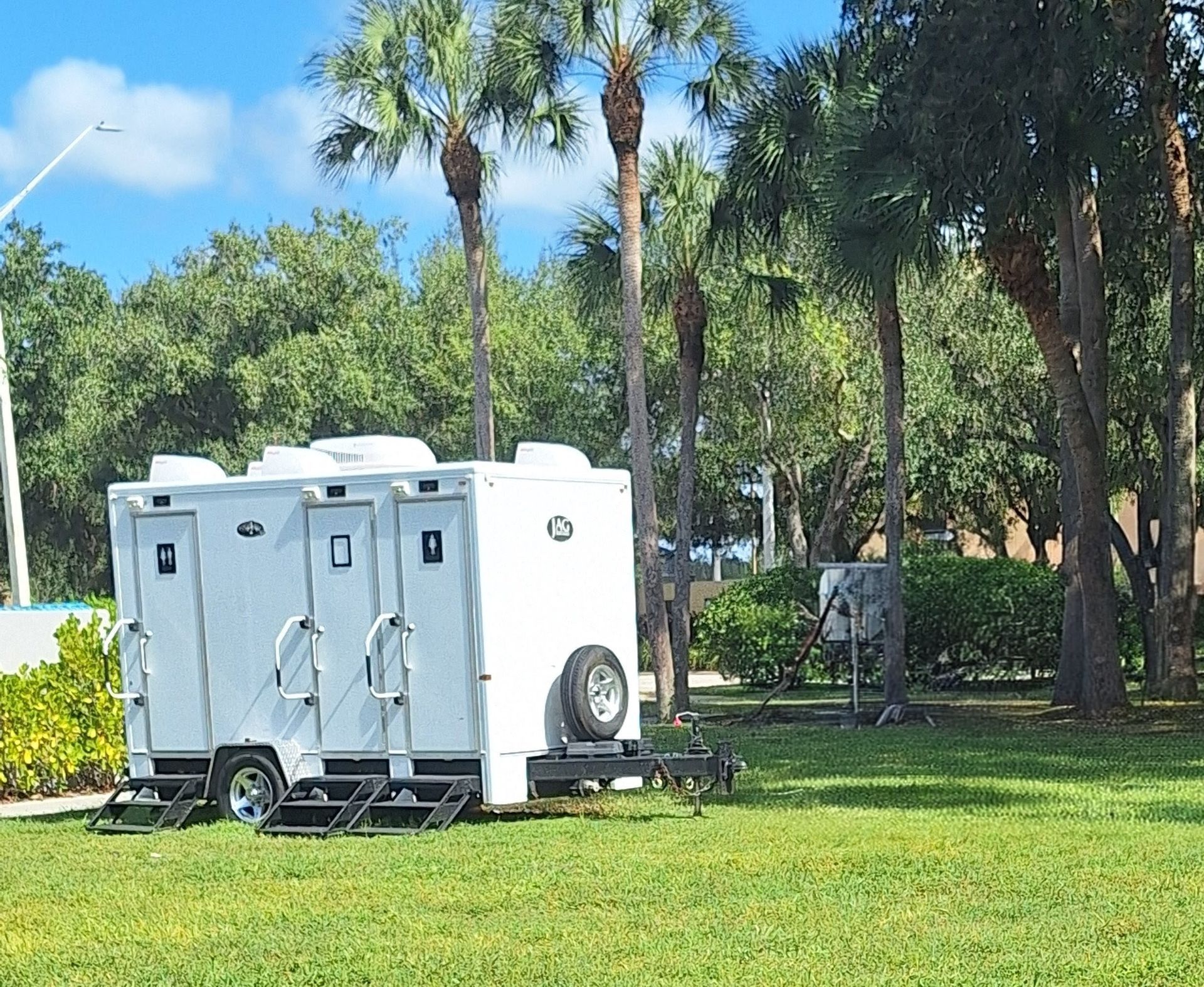 White portable restroom trailer on a grassy lawn with palm trees and blue sky behind it