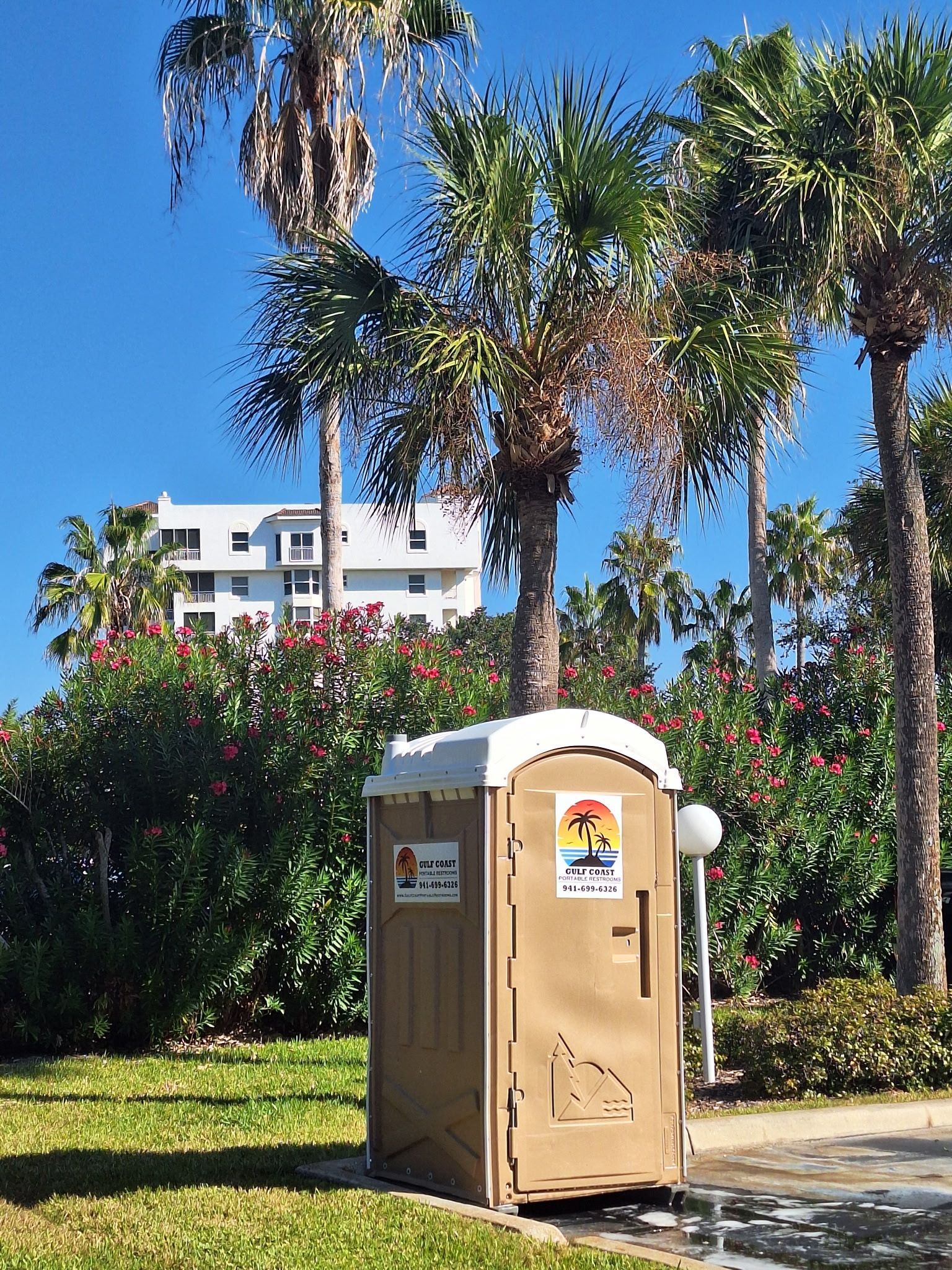 Portable toilet on a sunny lawn near palm trees and bushes, with a building in the background.
