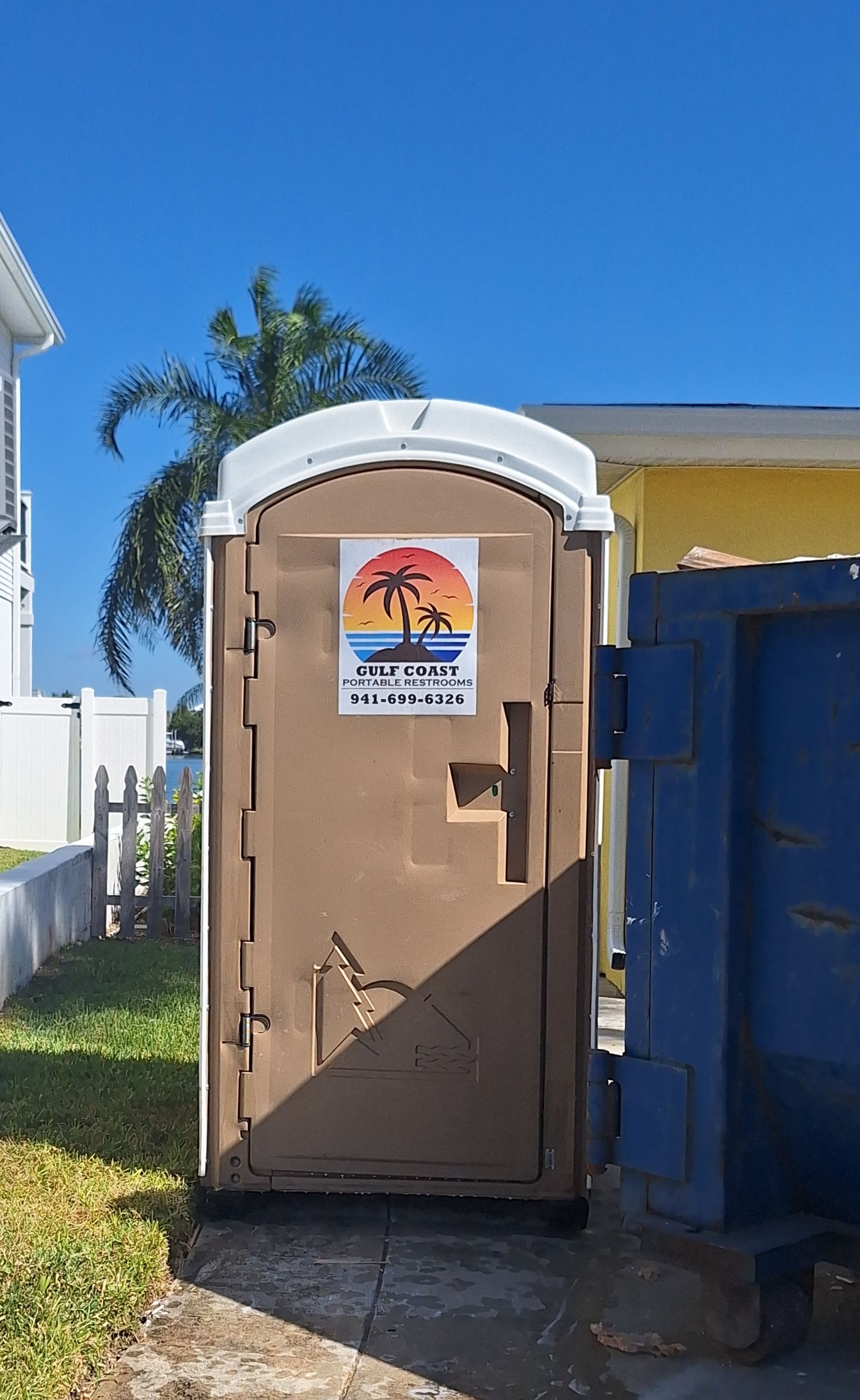 Portable toilet beside a blue dumpster on a sunny sidewalk, with a palm tree and fence behind it.