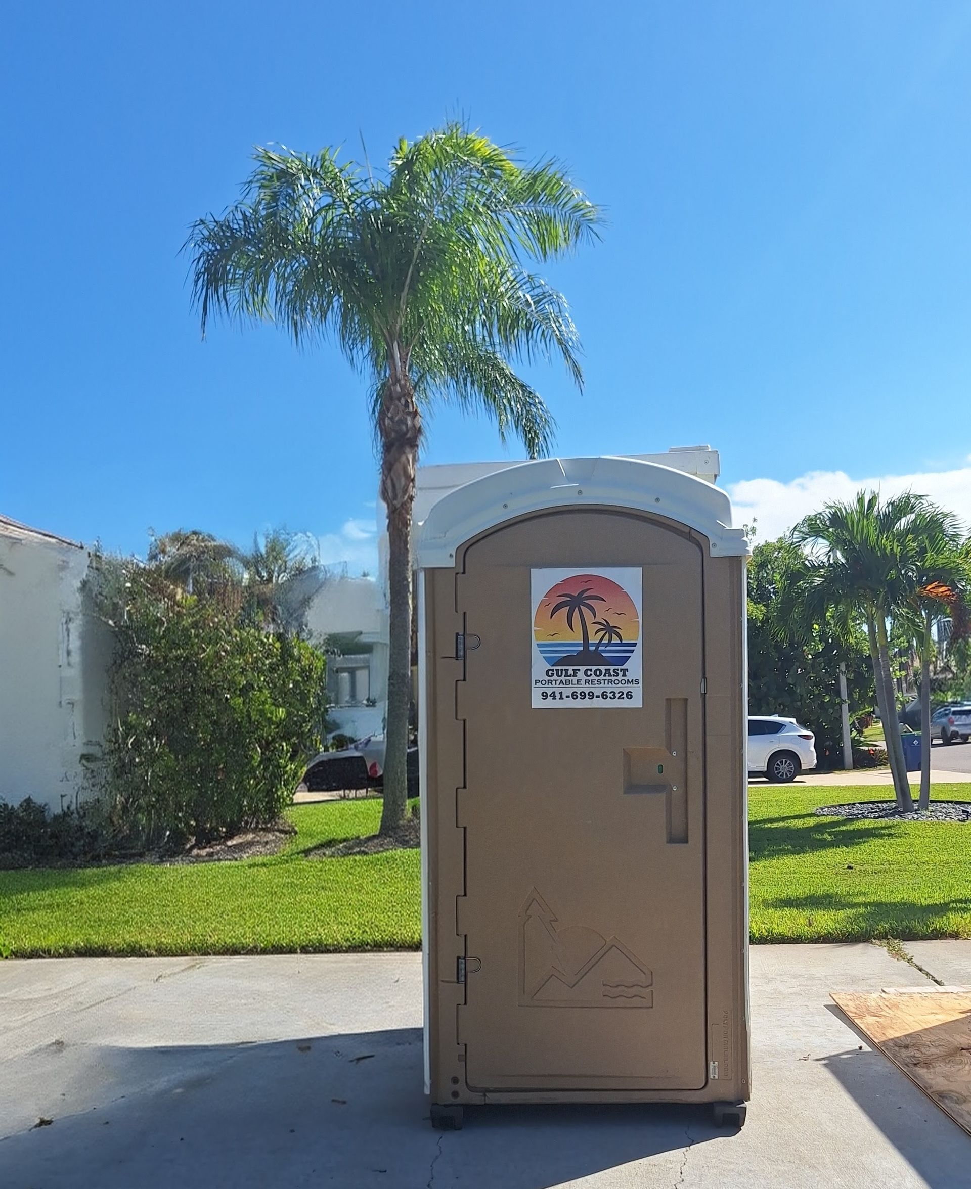 Portable toilet on a sunny street with a palm tree and houses in the background.