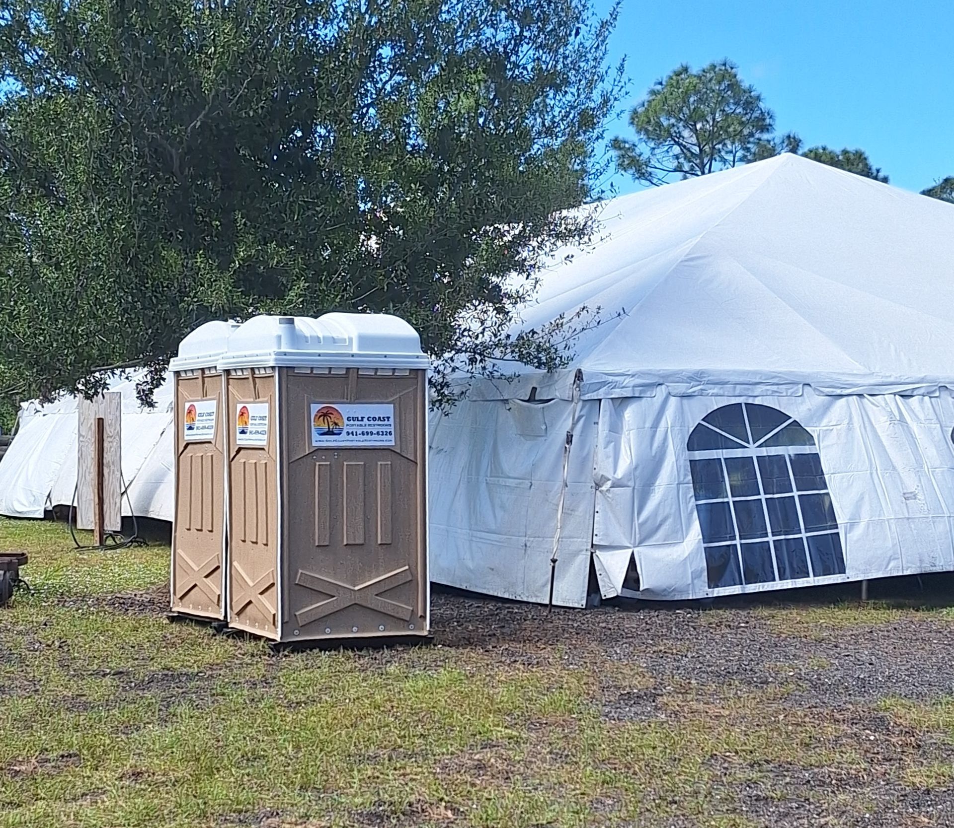 Portable toilet outside a large white event tent on a grassy field, with trees in the background