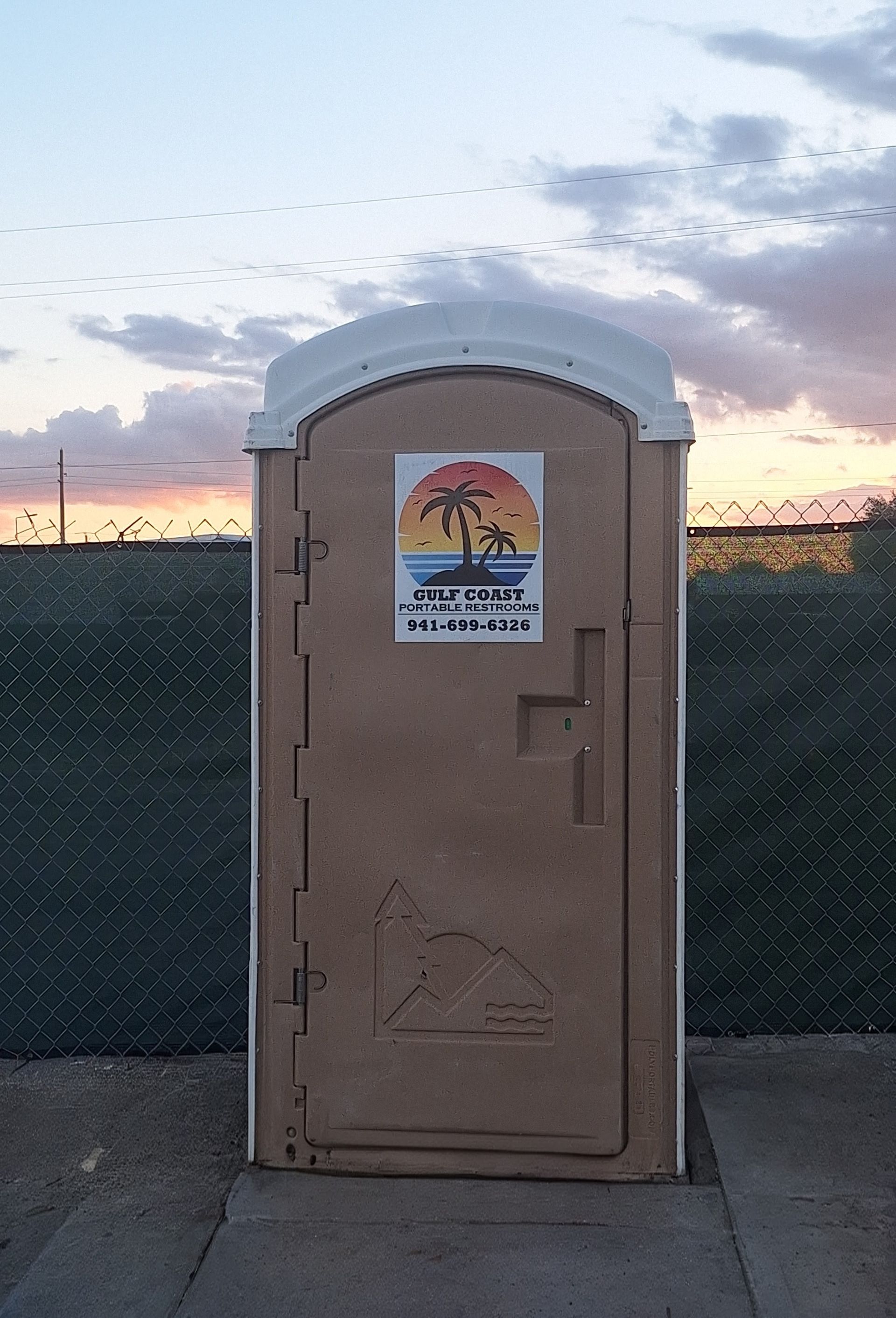 Portable toilet at dusk beside a fence, with a sunset sky in the background.