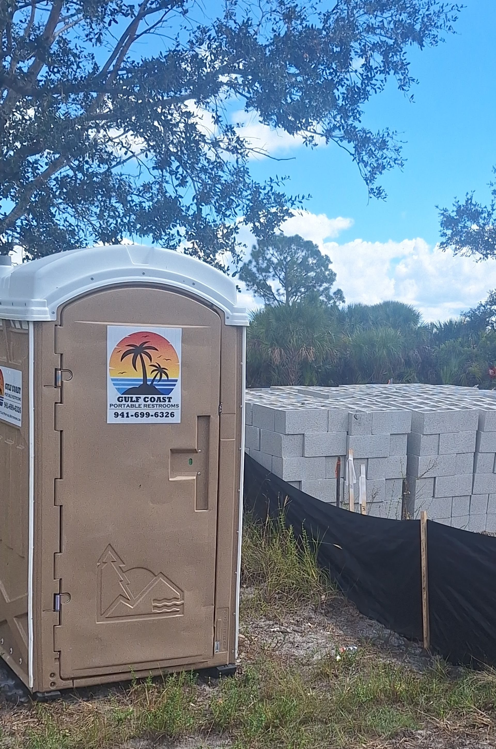 Tan portable toilet beside a white stone wall and trees under a blue sky.