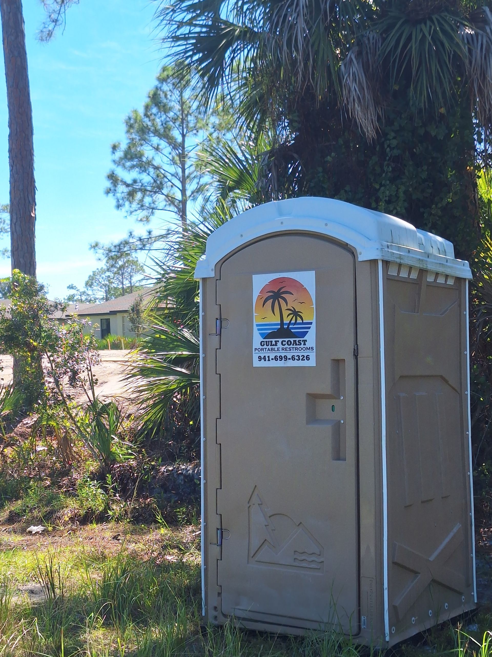 Tan portable toilet with snow on top in a sunny, wooded area