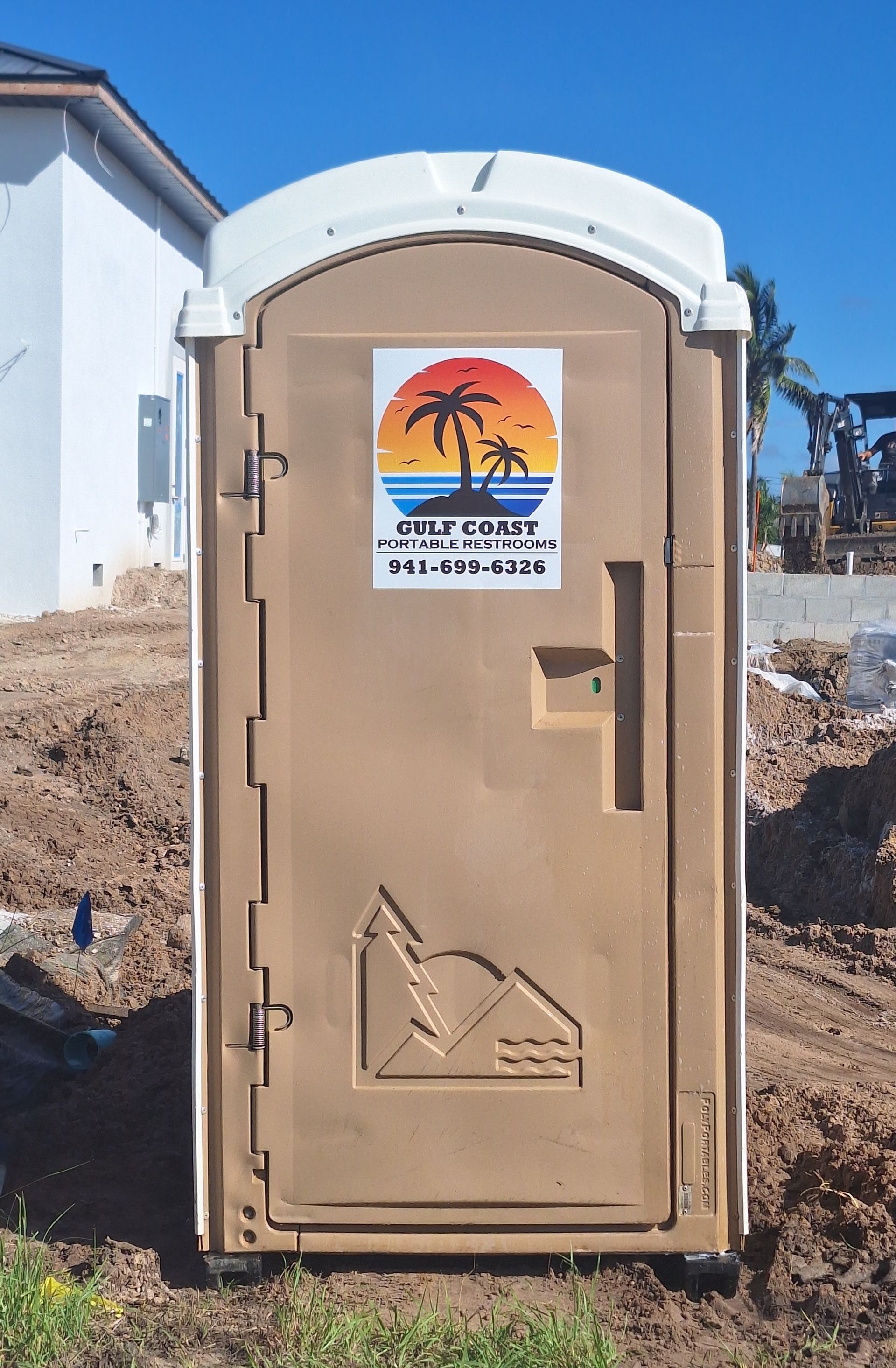 Portable toilet outside a building, with a sunset poster on the door and desert ground around it