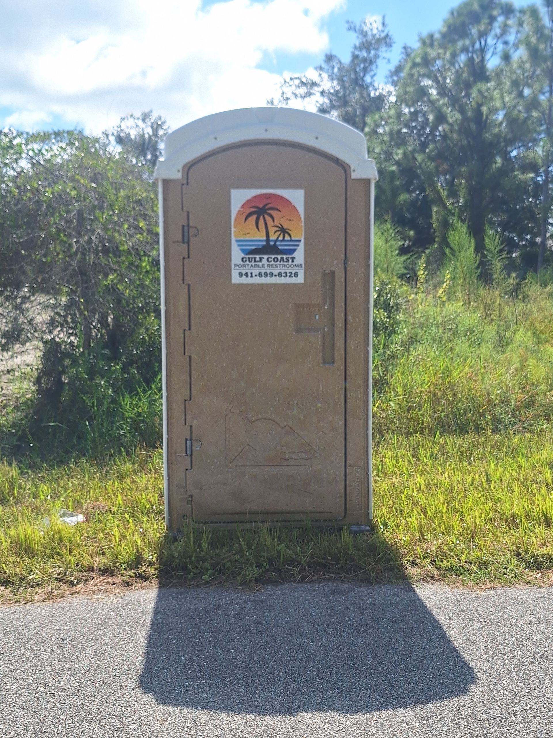 Portable toilet beside a path, with a warning sign and trees in the background.