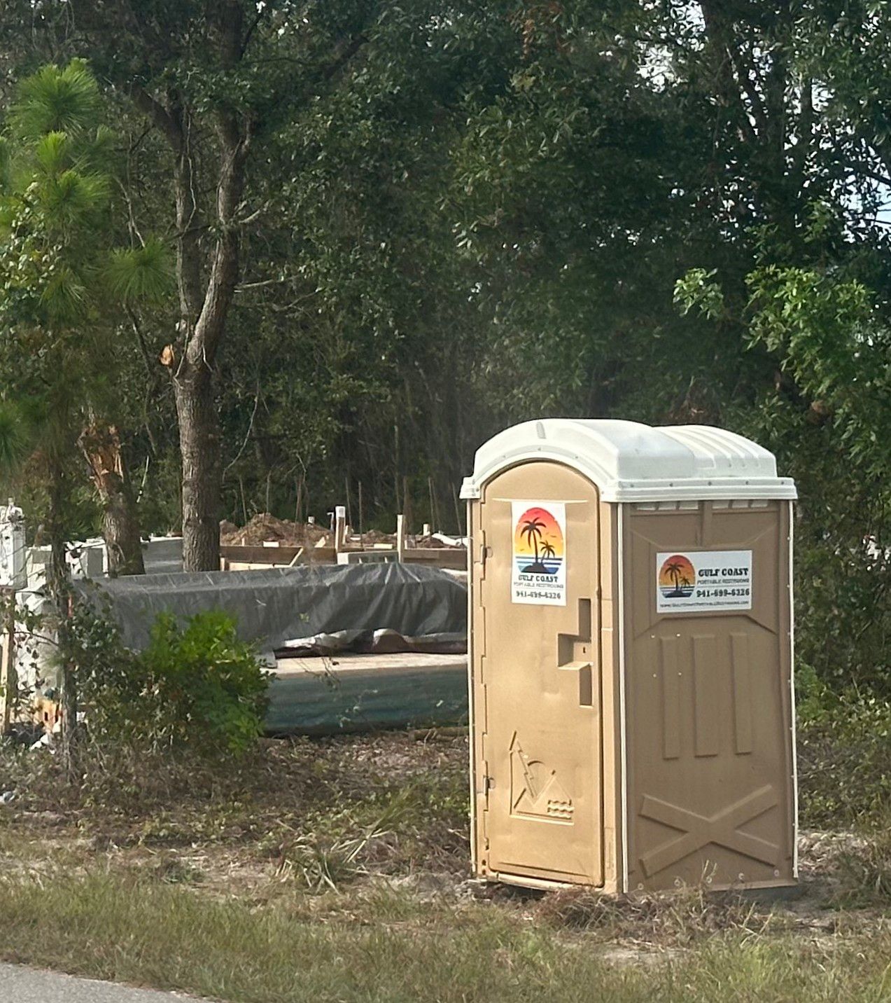 Beige portable toilet in a wooded park area with trees and a paved path in the background
