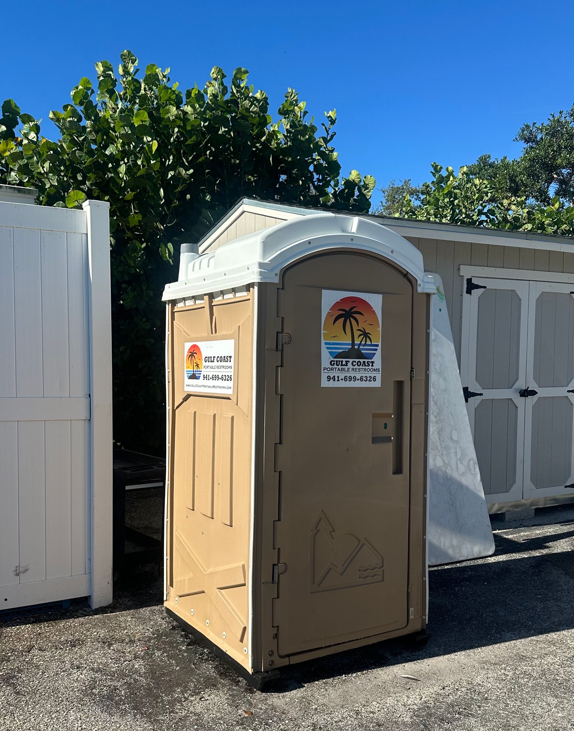 Portable tan portable toilet beside white fence under blue sky