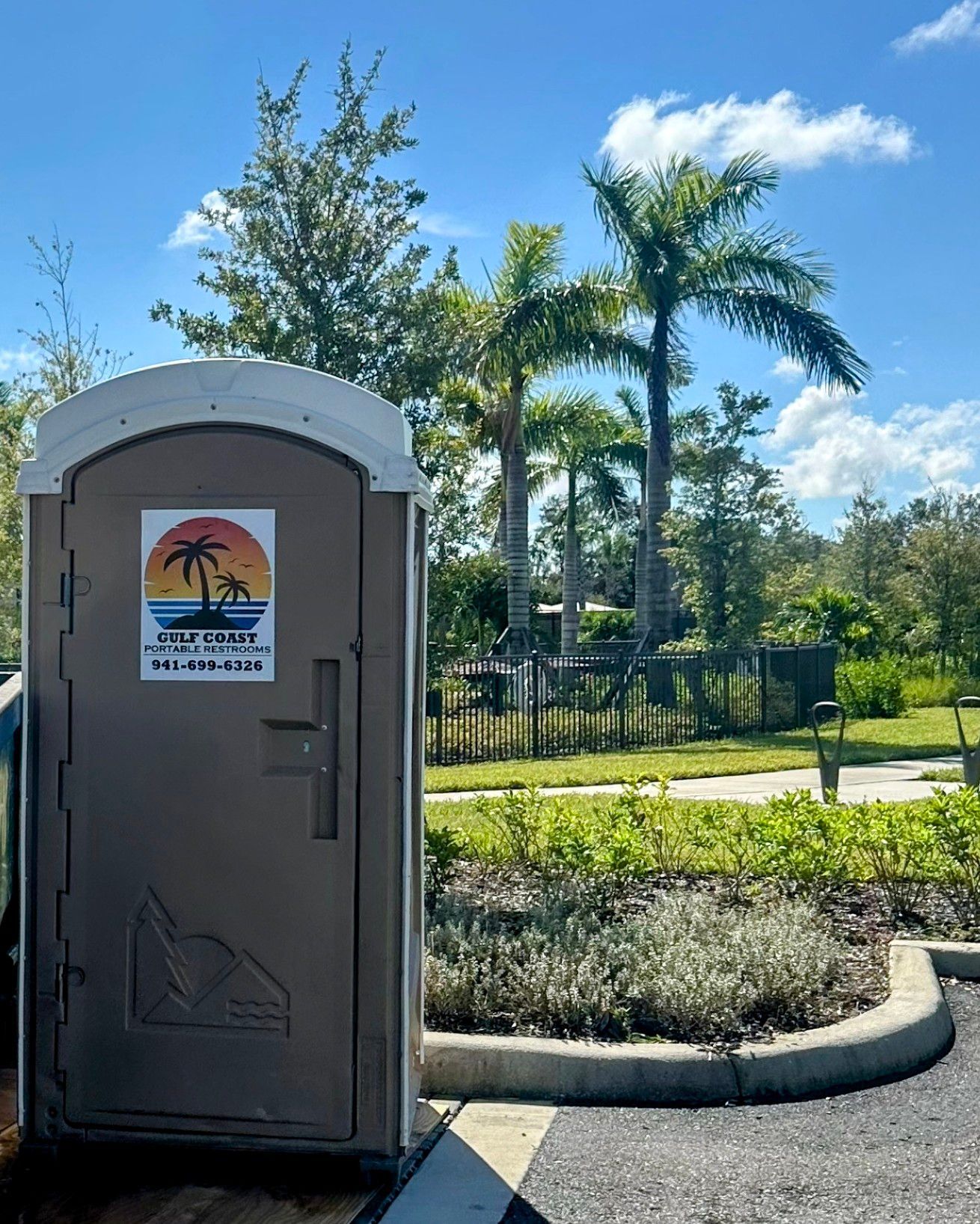 Portable restroom beside palm trees and fenced walkway under a bright blue sky.