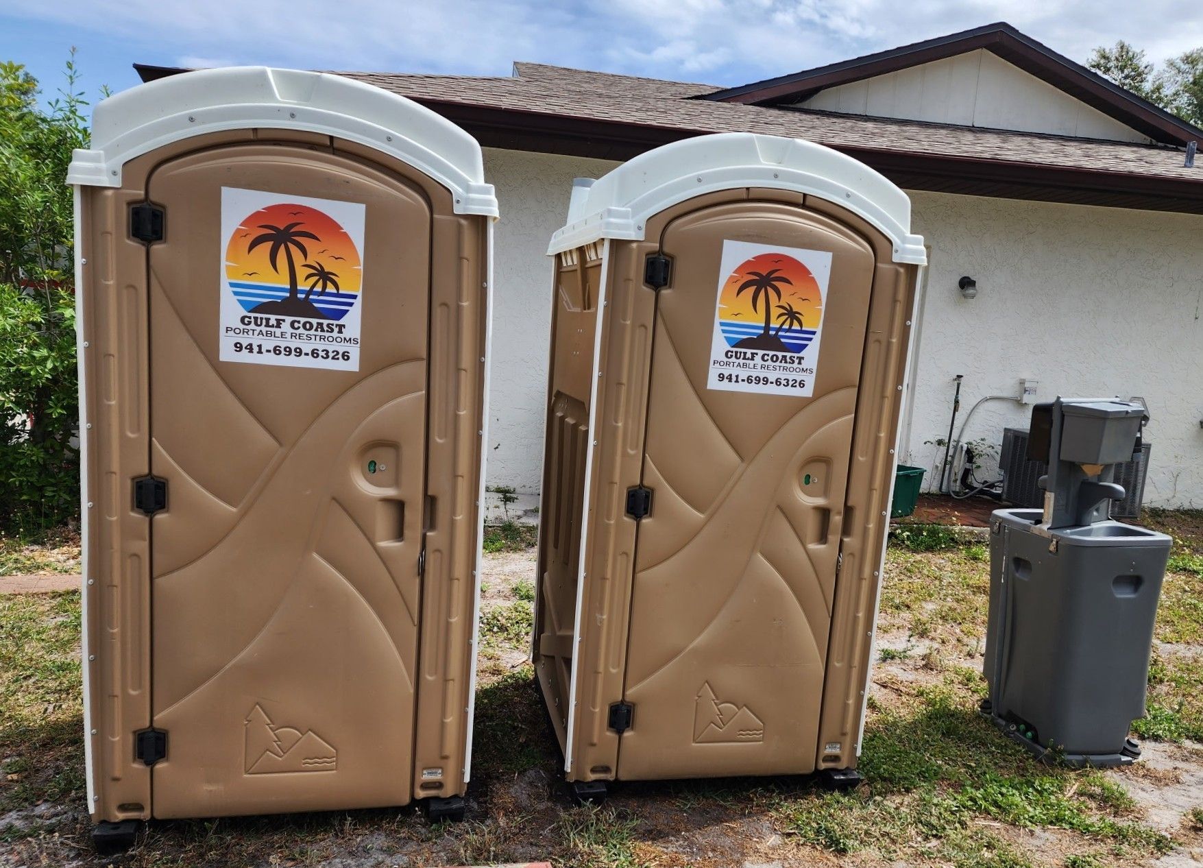 Two tan portable toilets beside a house, one with a trash bin and generator nearby.