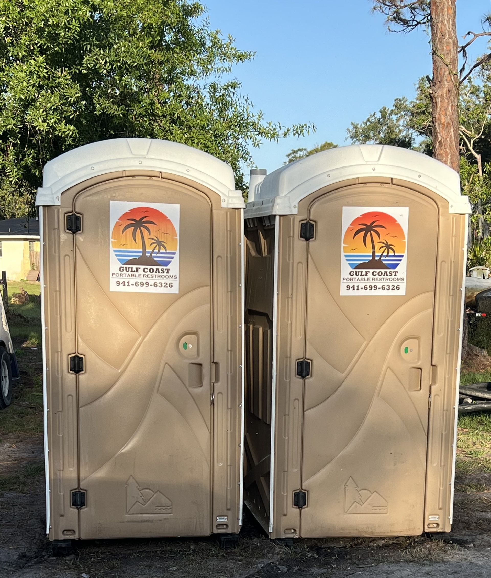 Two beige portable toilets side by side under trees, each with a sunset sign on the door.