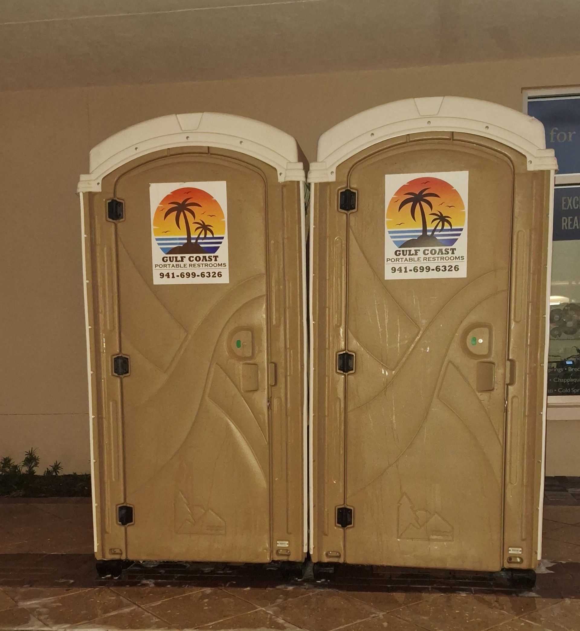 Two tan portable toilets with palm tree signs, standing side by side indoors.