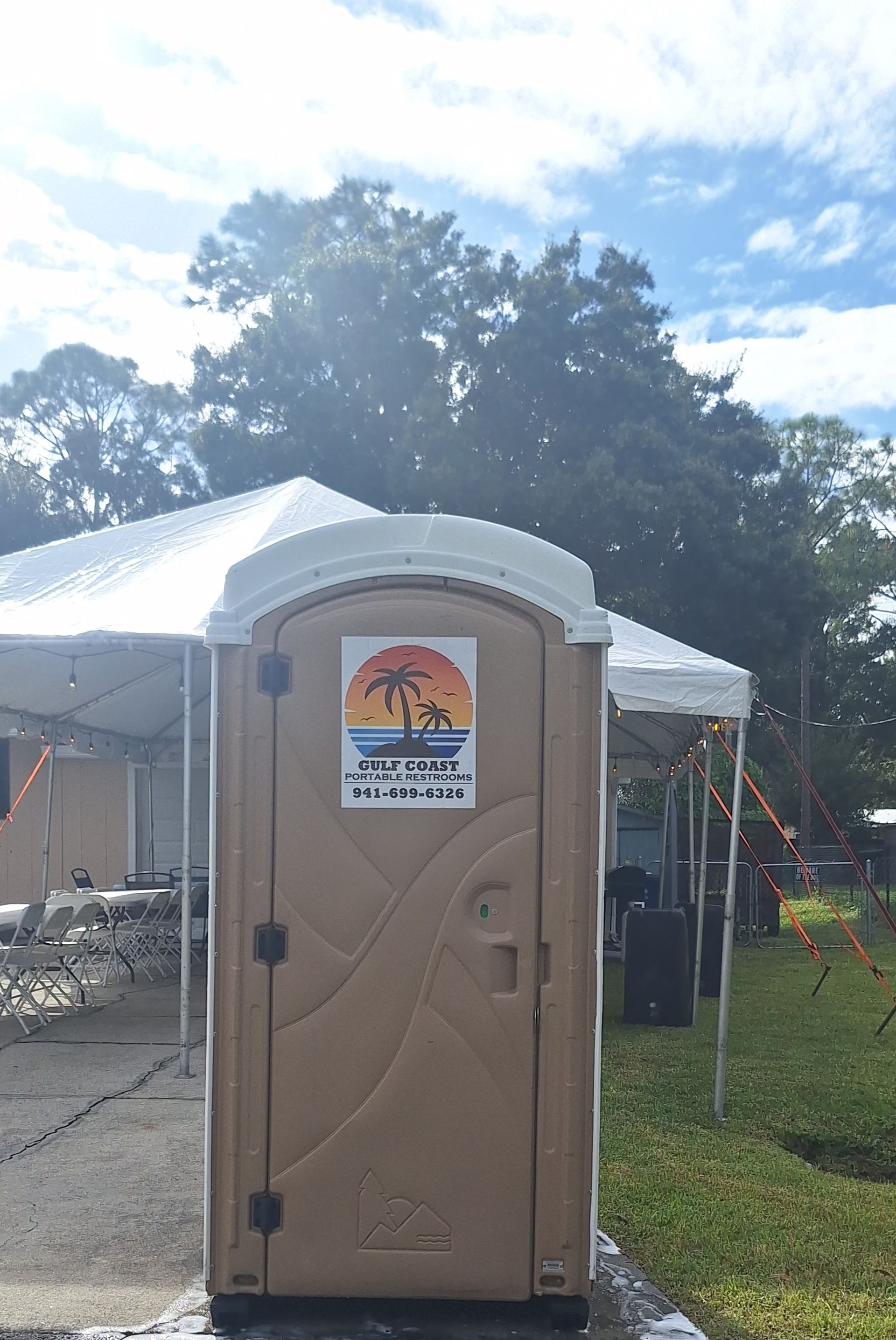 Tan portable toilet beside white tents on a grassy outdoor event site under a bright sky