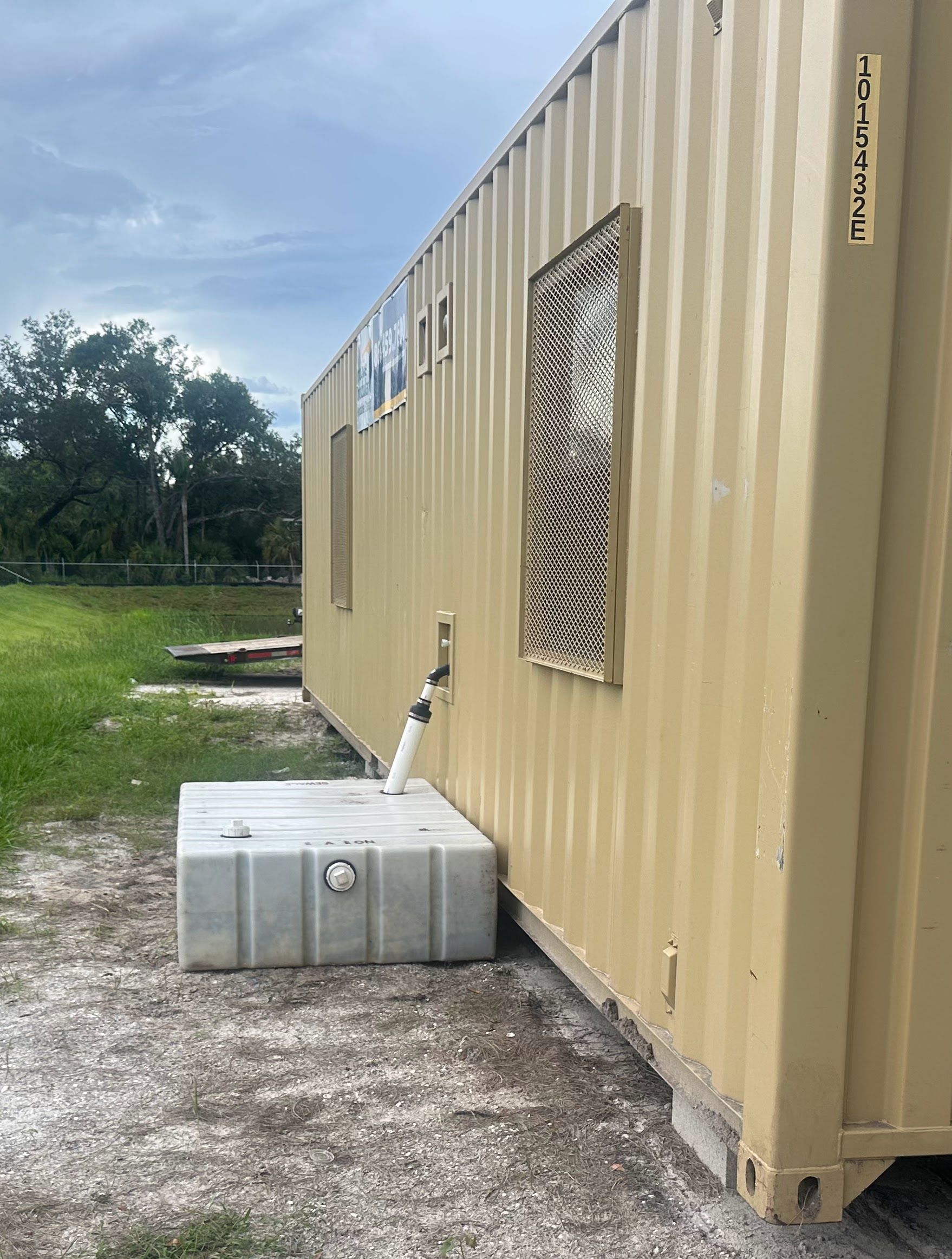 Beige shipping container with barred windows and a small white tank on gravel beside a grassy field.