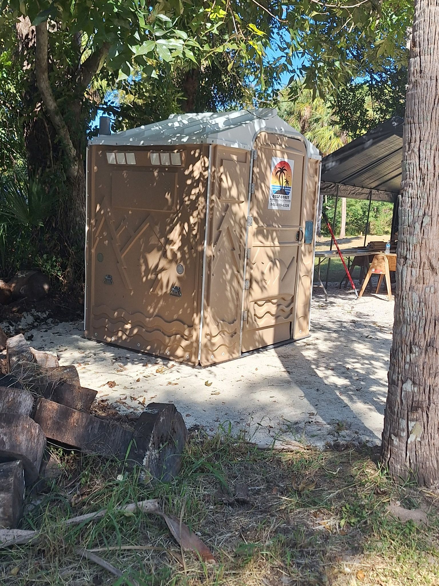 Brown portable toilet in a shaded wooded area, with a concrete path and trees around it