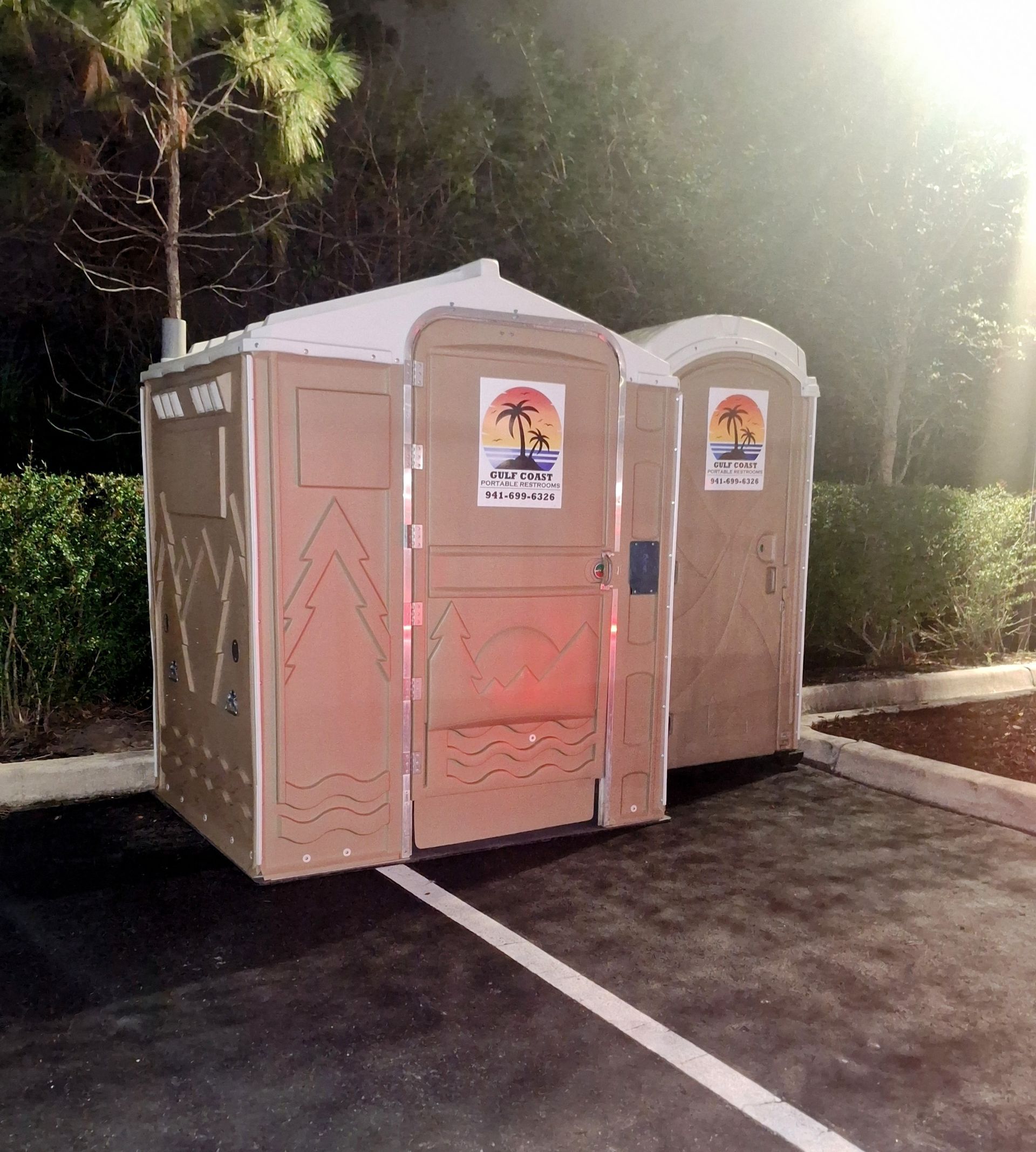 Two beige portable toilets in a parking lot, one door open, with trees and sunlight behind them