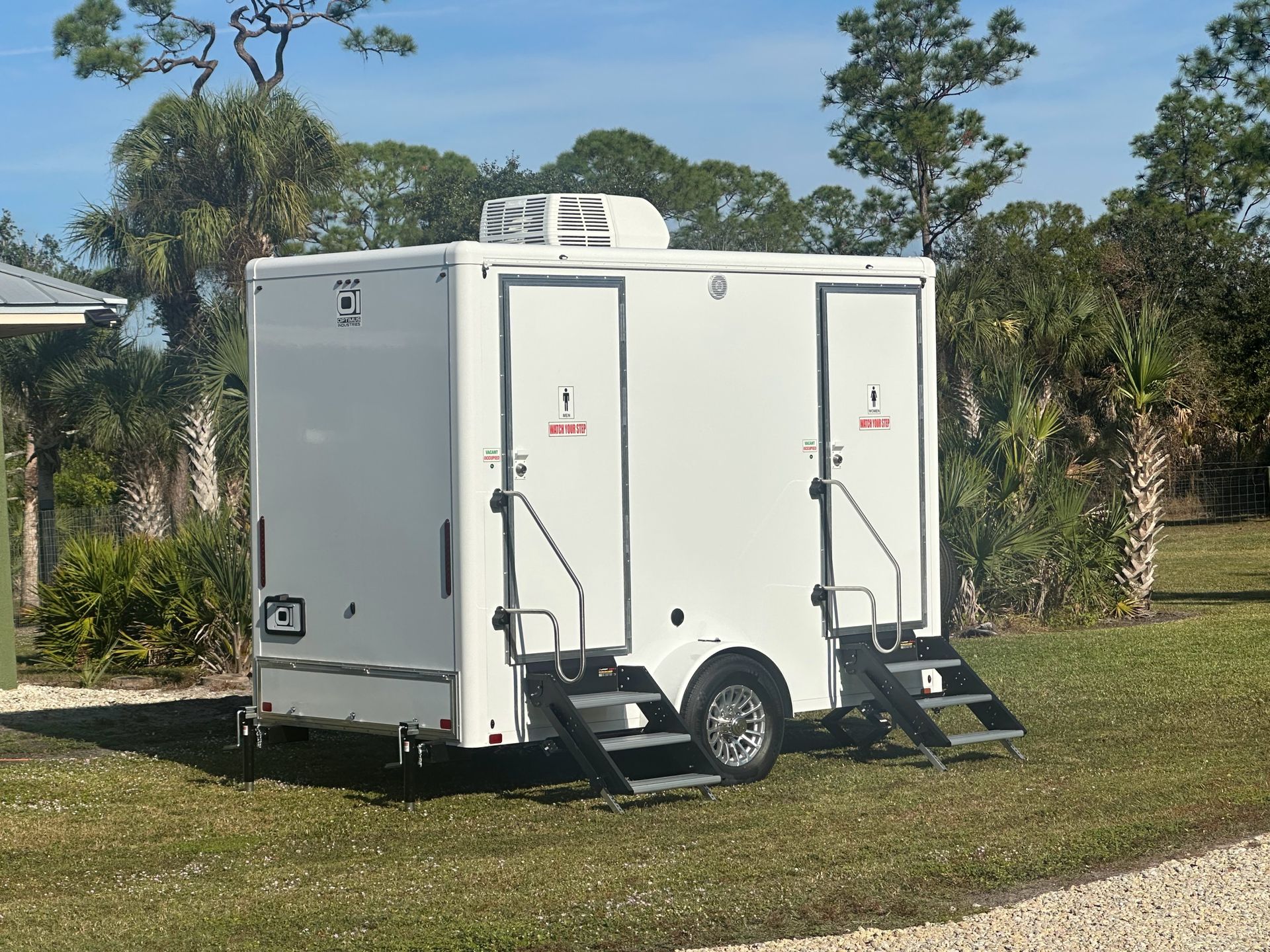 White portable toilet trailer with two access ramps parked on grass outdoors.