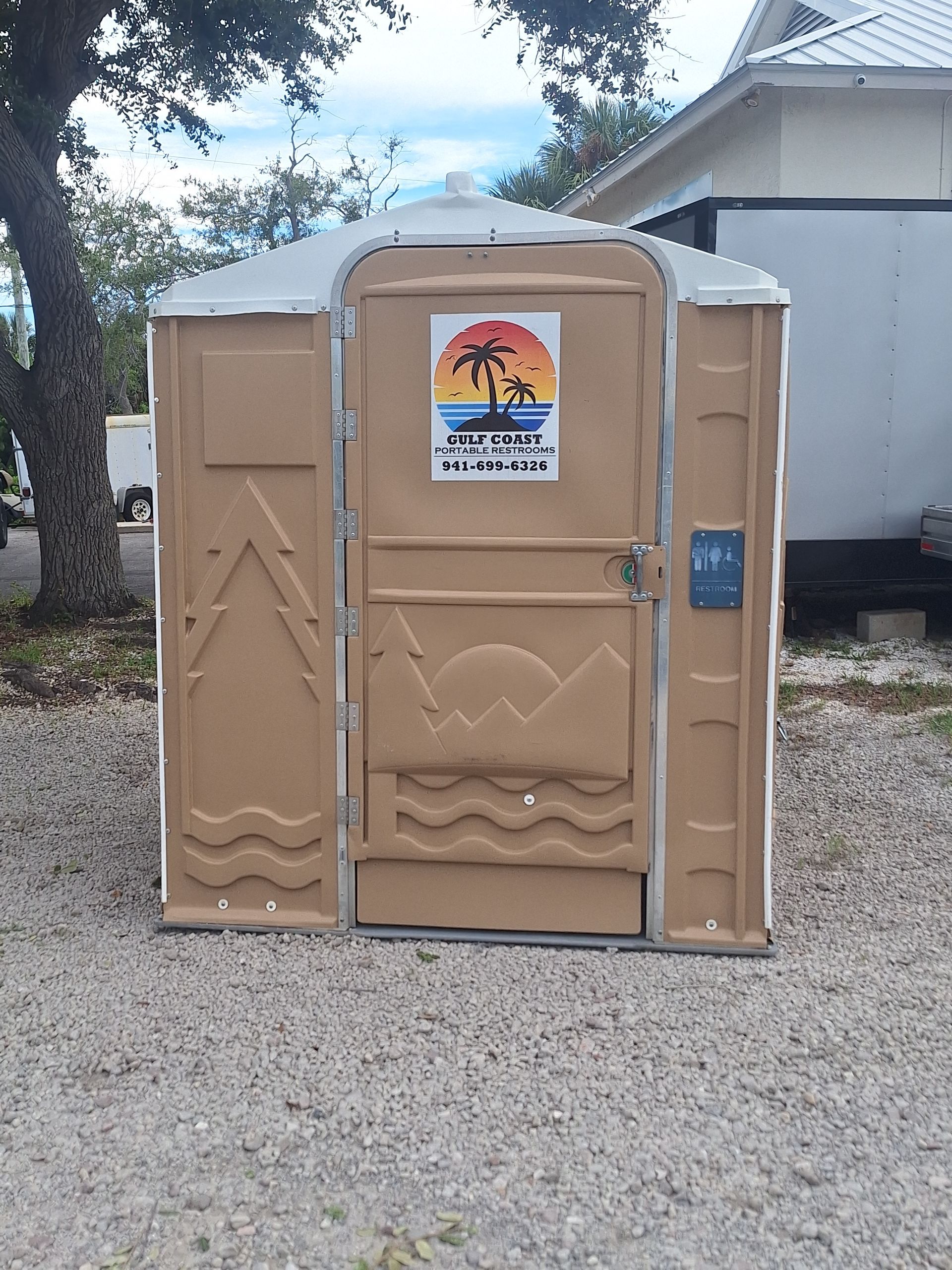 Tan portable toilet on gravel beside a house, with a colorful sign on the door.