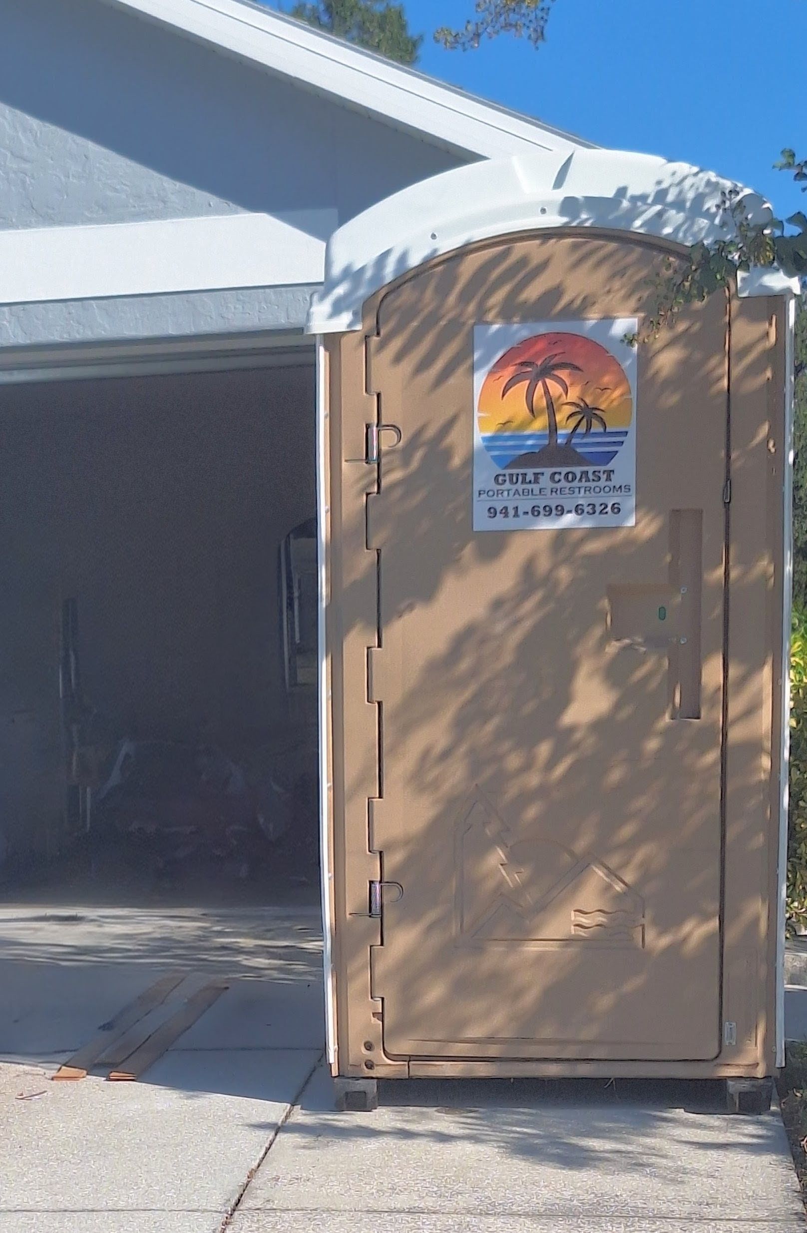 Tan portable toilet beside a garage, with a rainbow sign on the door.