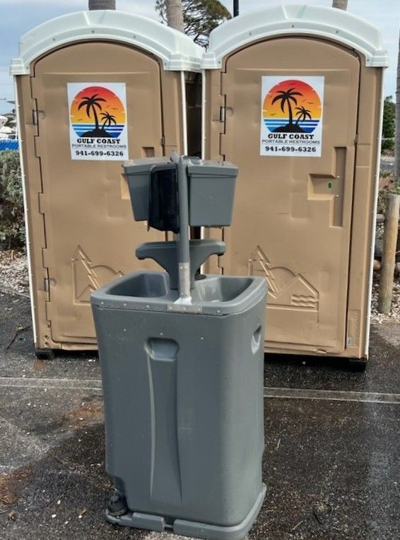 Gray handwashing station in front of two portable toilets with signs attached.