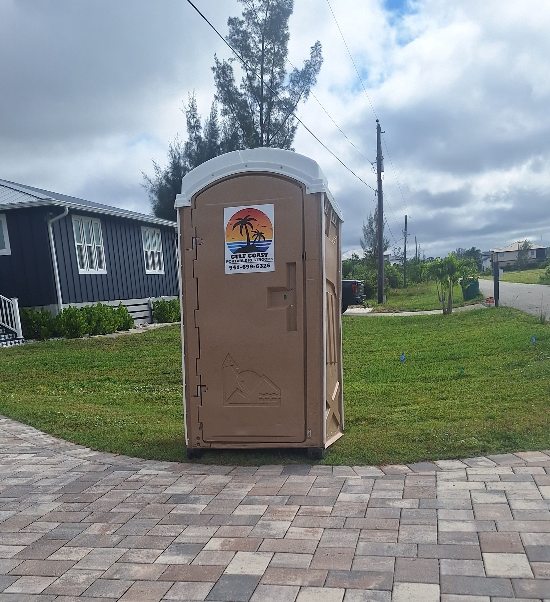Portable toilet on a paved lawn beside a road under a cloudy sky.
